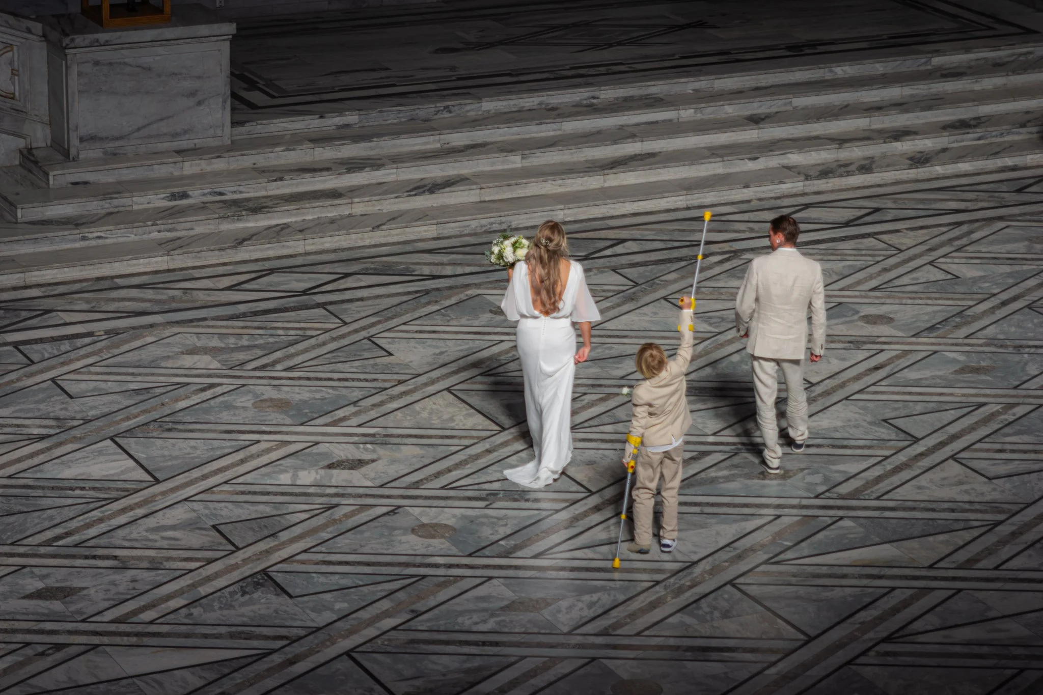 A bride and groom walking down a marble hall with two children, one holding a bouquet of flowers. The children are dressed in light-colored clothing and yellow and white bowling pins are seen on the floor, suggesting a playful or unconventional weddi