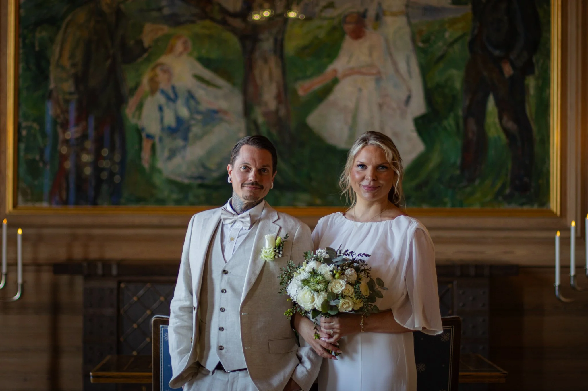 A couple dressed in wedding attire standing inside a church with a large painting of a biblical scene in the background. The man wears a cream-colored suit with a bow tie, and the woman wears a white wedding dress holding a bouquet of white and purpl
