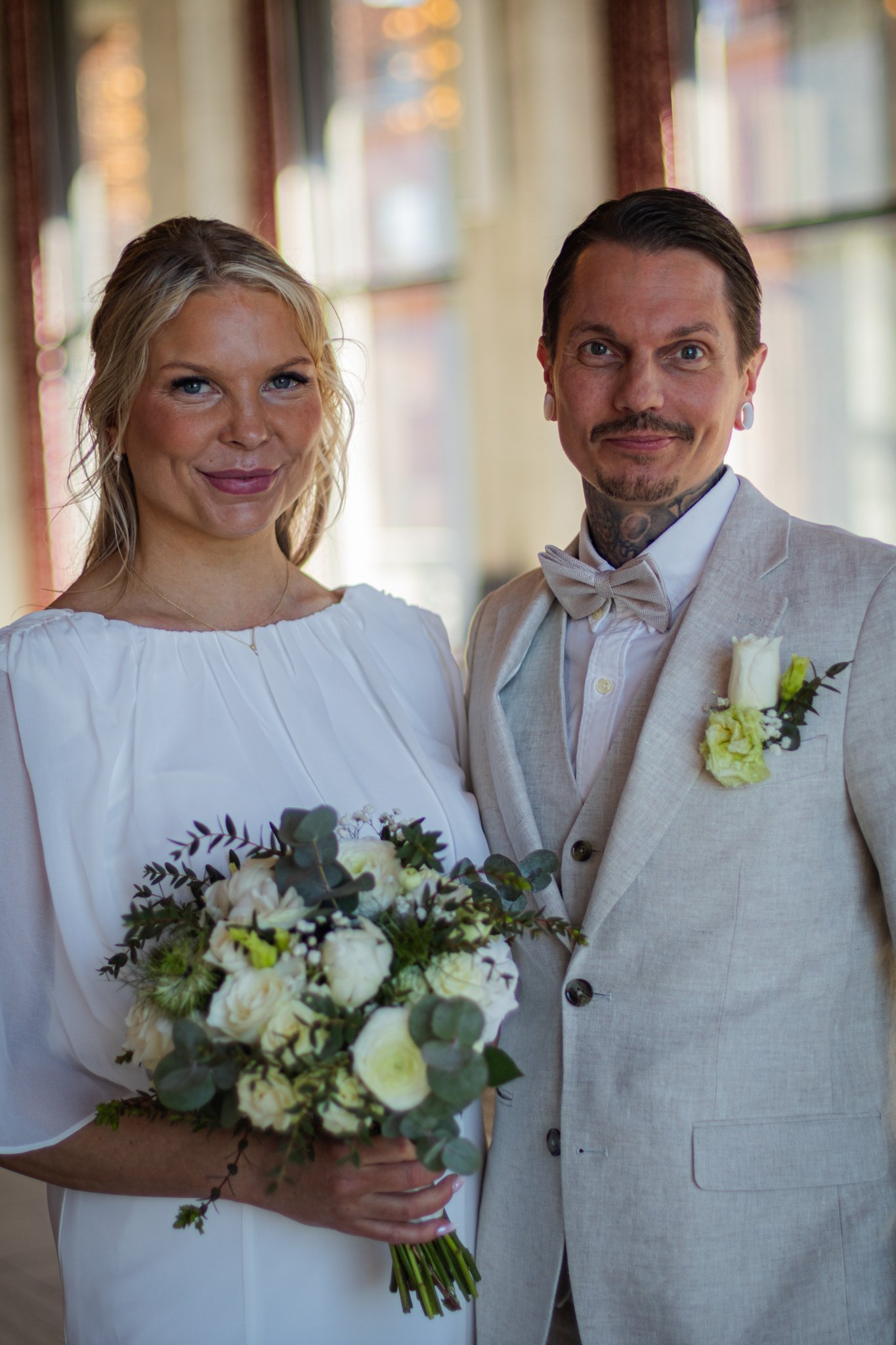 A bride and groom dressed in wedding attire standing together indoors, with the bride holding a bouquet of white flowers and greenery.