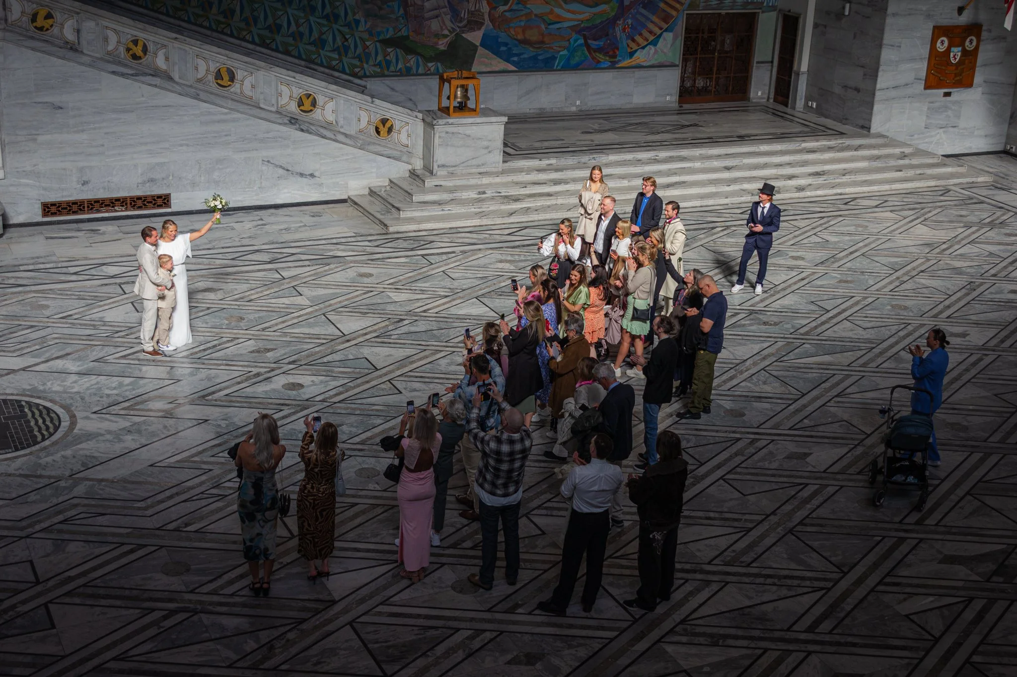 A wedding ceremony taking place inside a grand marble hall, with a bride and groom standing together with young children, surrounded by an audience taking pictures and watching.