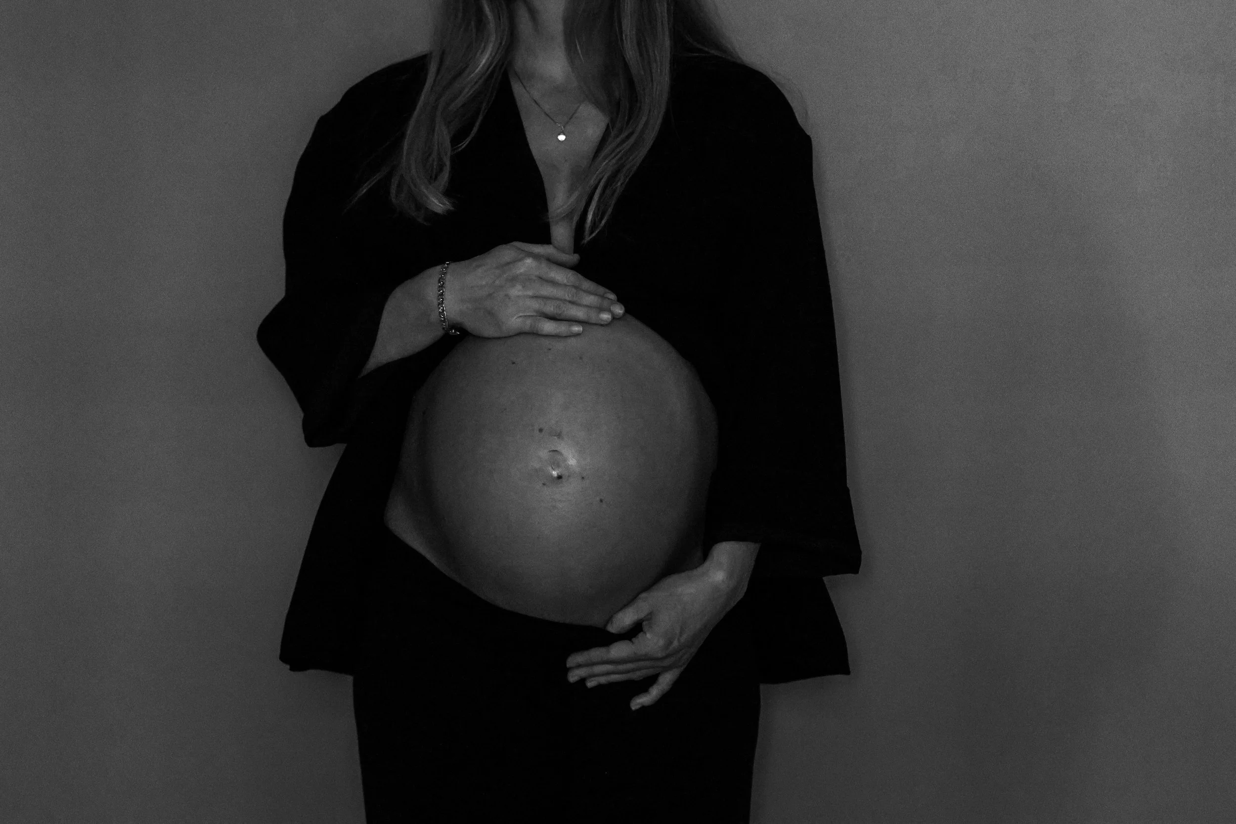 A woman with long hair is holding her pregnant belly. She is wearing a dark blazer and jewelry, and the background is plain.