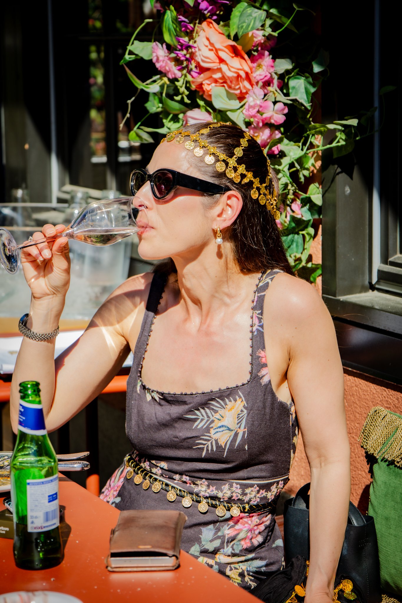 A woman with long dark hair, wearing sunglasses, gold coin head jewelry, earrings with pearls, and a floral dress, is drinking from a wine glass. She is sitting at an orange table with a bottle of beer and a wallet in front of her. In the background,