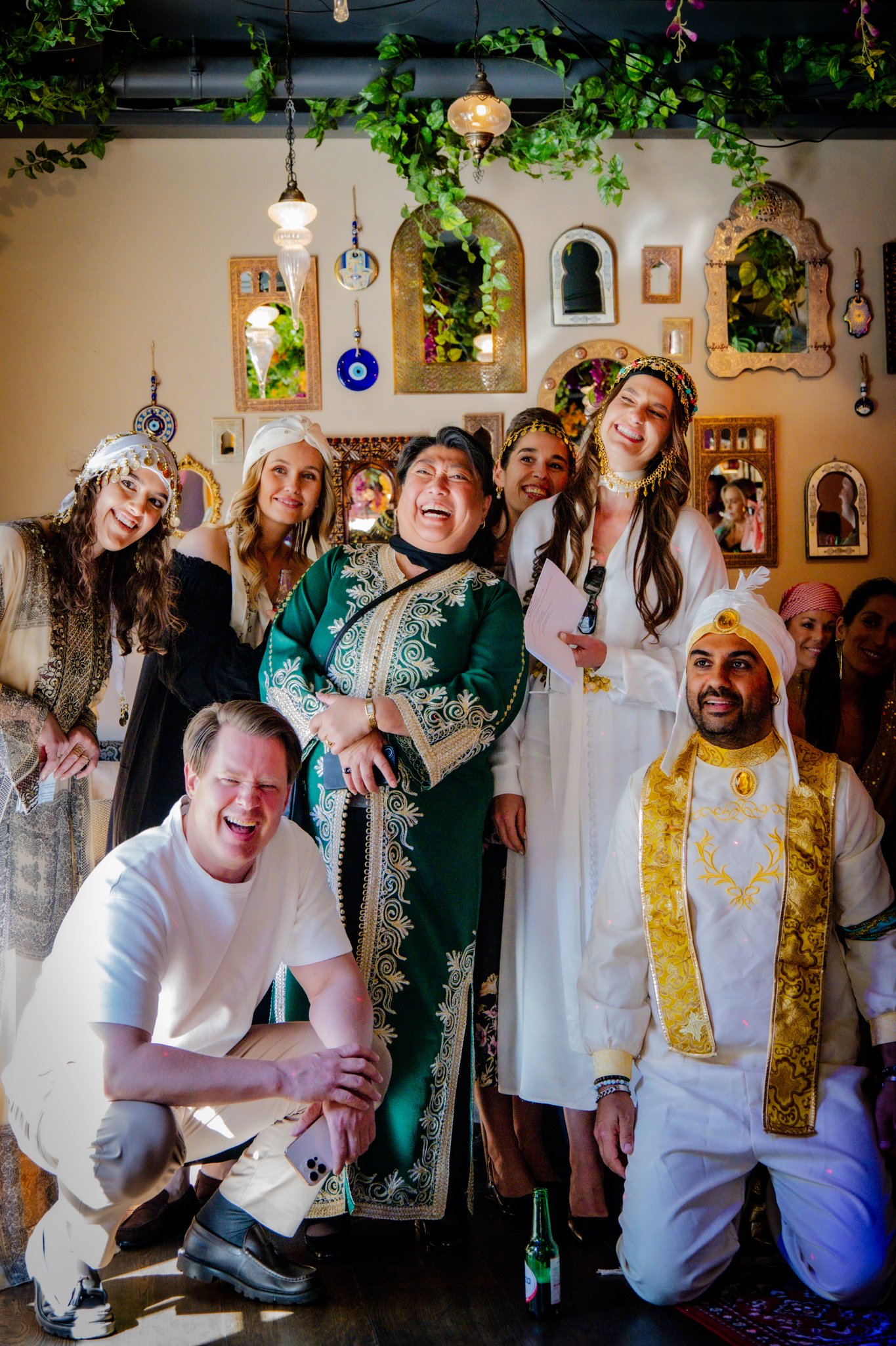 A diverse group of people dressed in traditional and cultural attire, gathered indoors with decorative mirrors and hanging lights, smiling, and posing for the photo.