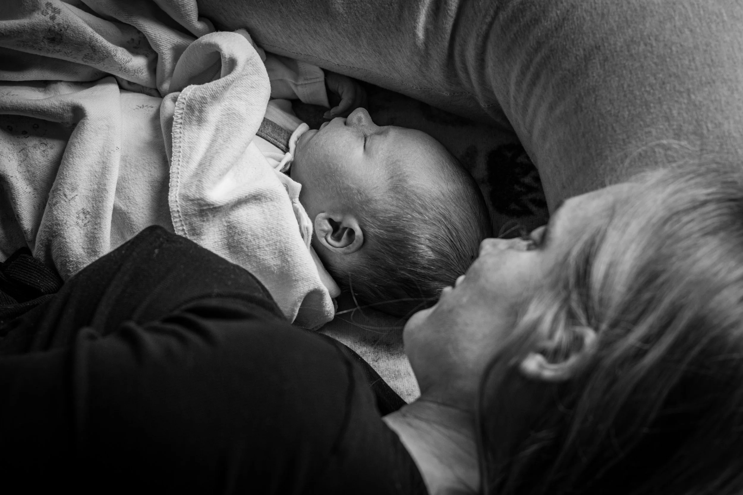 A sleeping baby with light hair, wrapped in a towel, resting on an adult's lap or chest, with the adult wearing a long-sleeved shirt or sweater. The scene is in black and white.