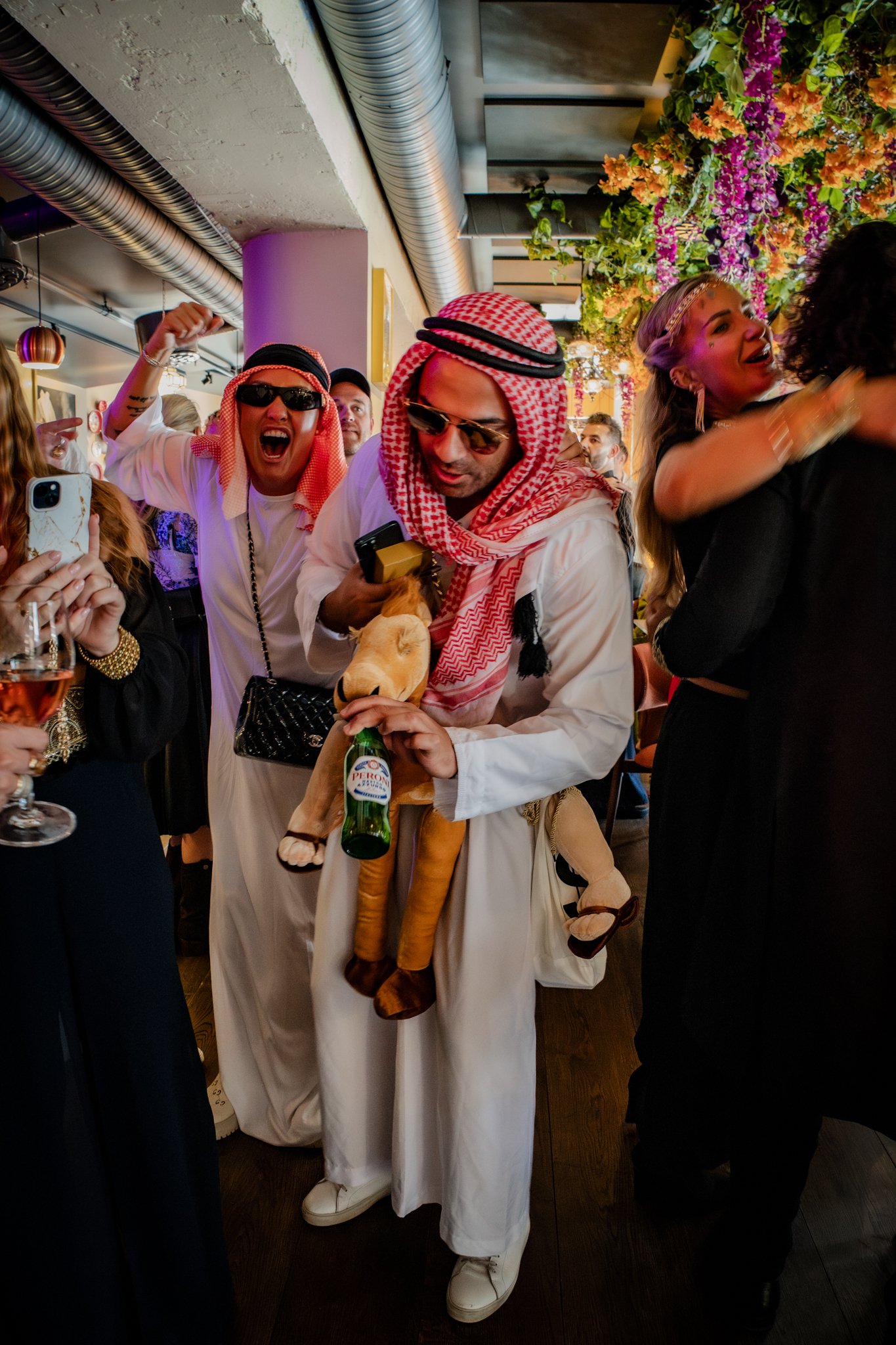 Group of people celebrating in a lively indoor setting, with two men wearing traditional Middle Eastern attire, including red and white checkered keffiyeh scarves, holding a beer bottle. Other guests are seen smiling, hugging, and taking photos, surr