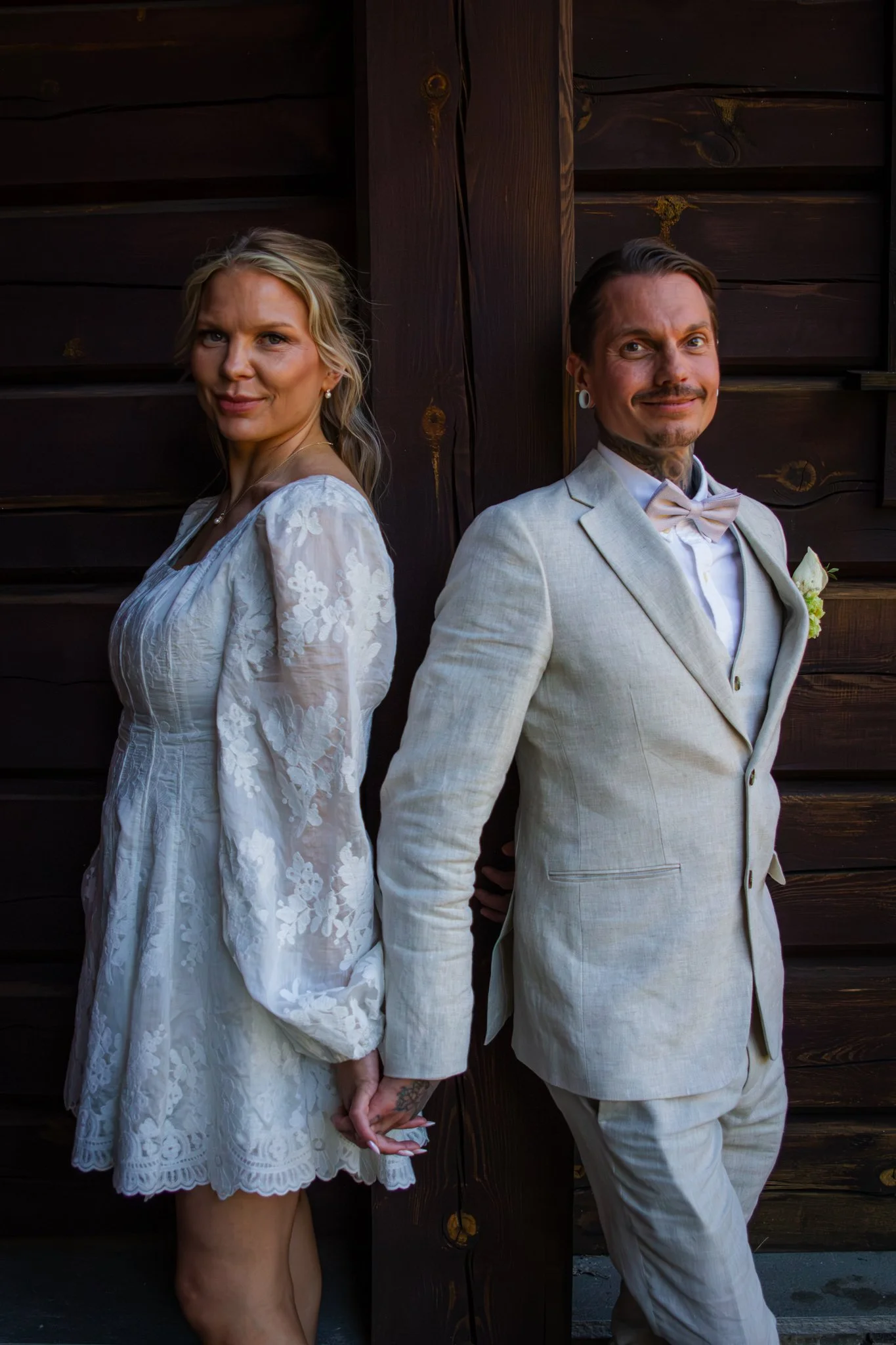 A man and woman standing back to back in front of a wooden wall, holding hands, dressed in wedding attire, with serious expressions.