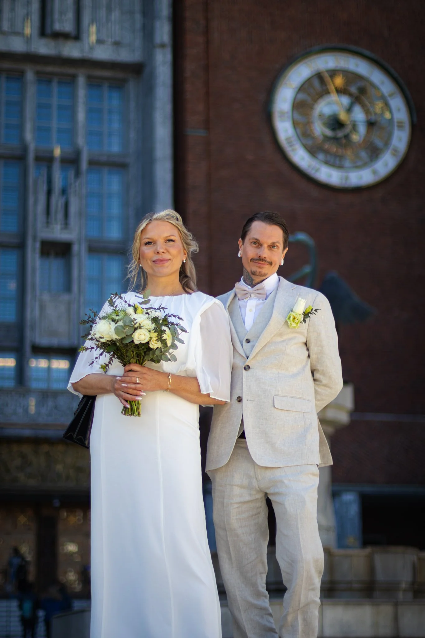 A bride and groom standing outdoors in front of a building with a clock tower. The bride is holding a bouquet of white flowers and wearing a white dress with puffed sleeves. The groom is wearing a light-colored suit with a bow tie and boutonniere.