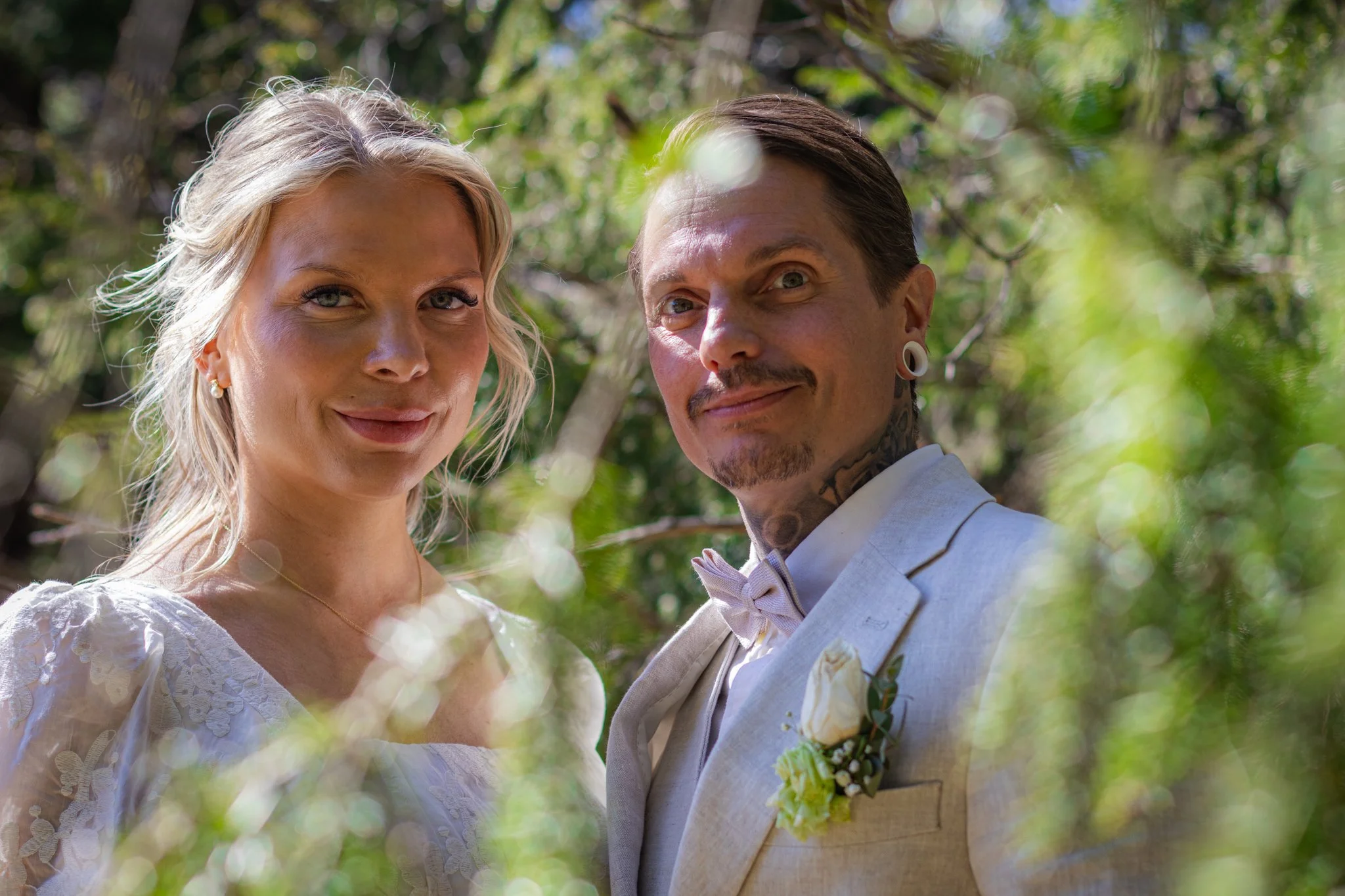 A couple dressed in wedding attire, standing outdoors among trees, smiling and looking at the camera.