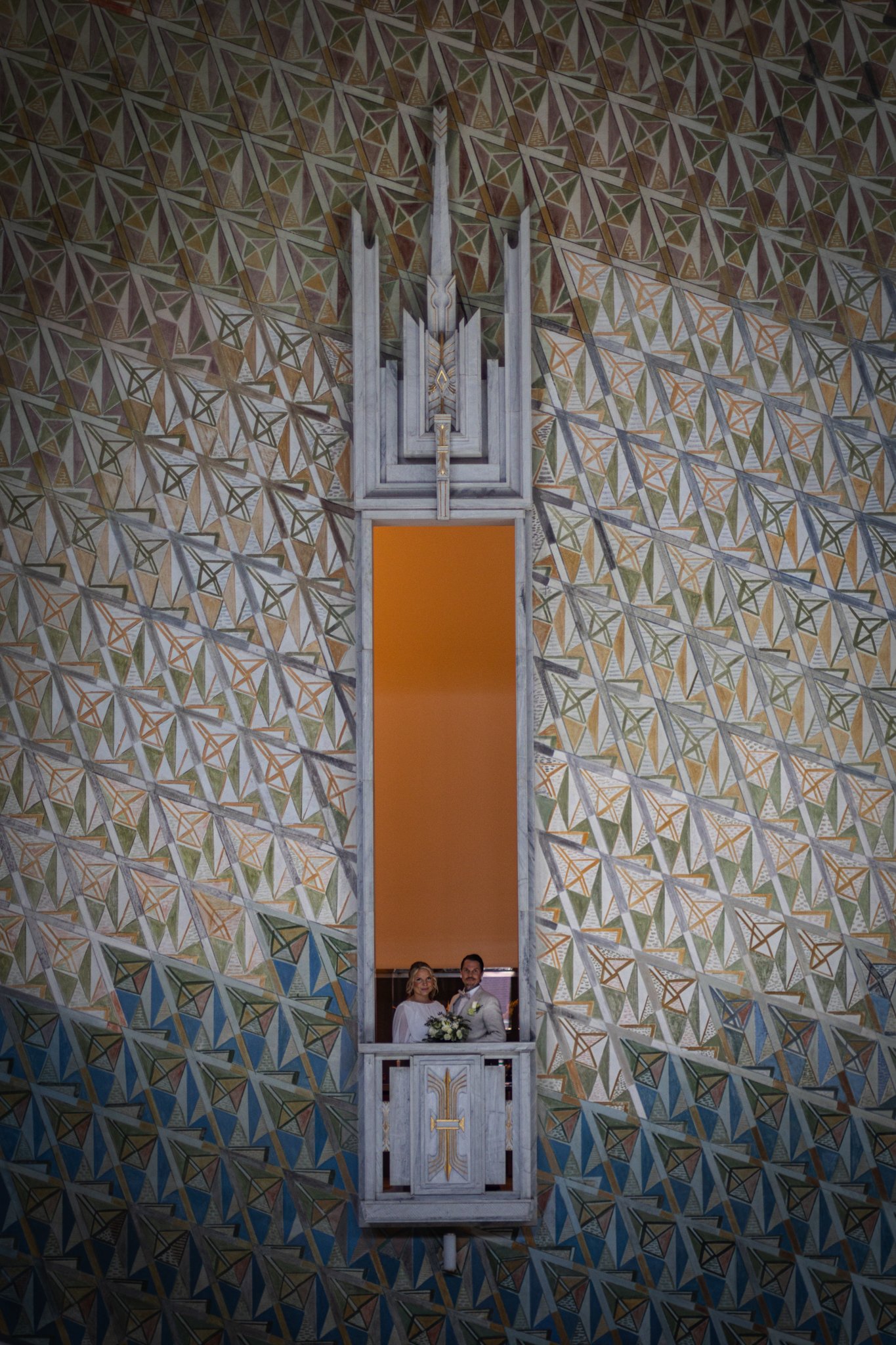 A bride and groom standing in a small balcony with a geometric patterned wall behind them.