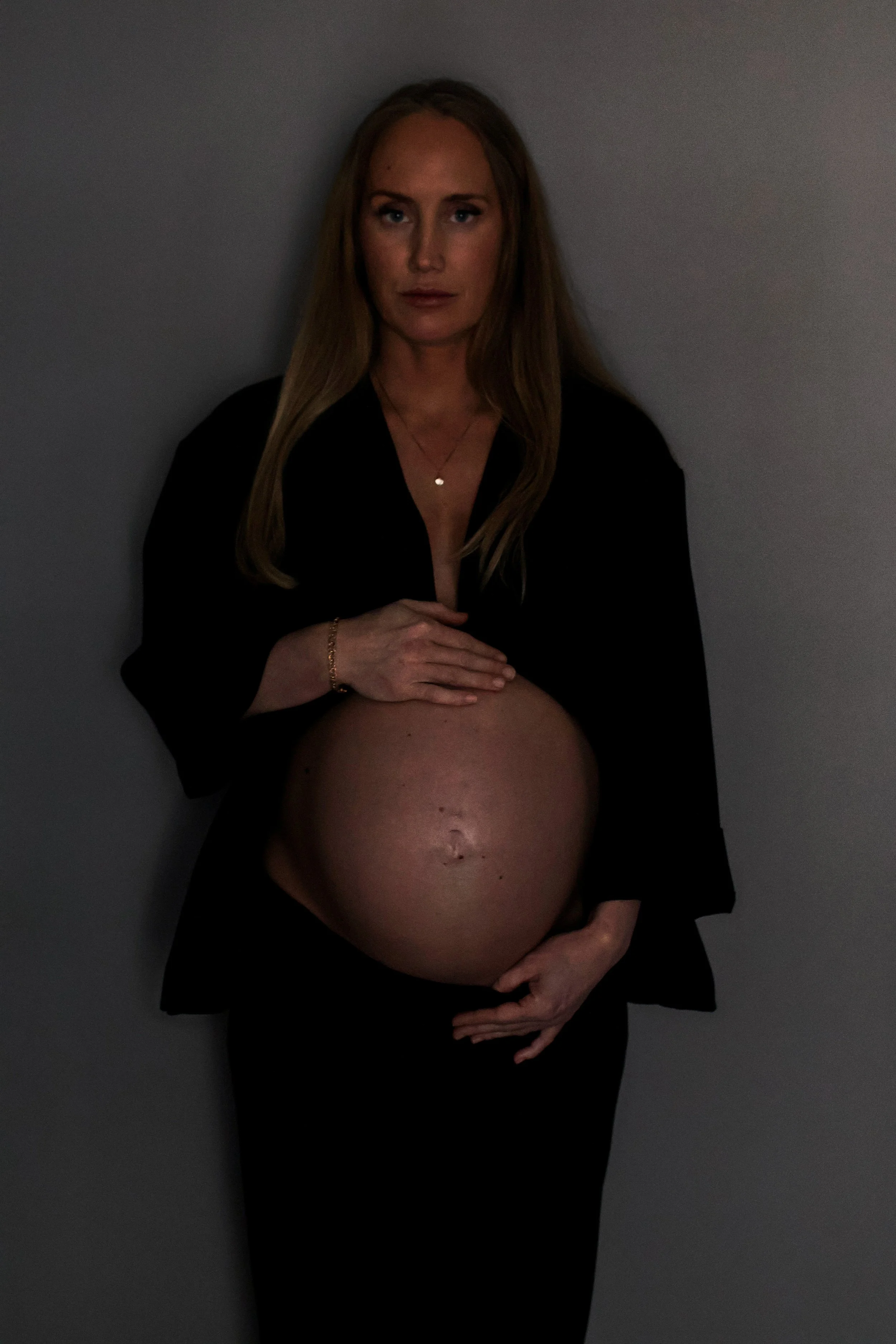 A woman with long blond hair, wearing a black outfit, holding her pregnant belly with both hands. She is standing against a plain gray wall, looking directly at the camera.