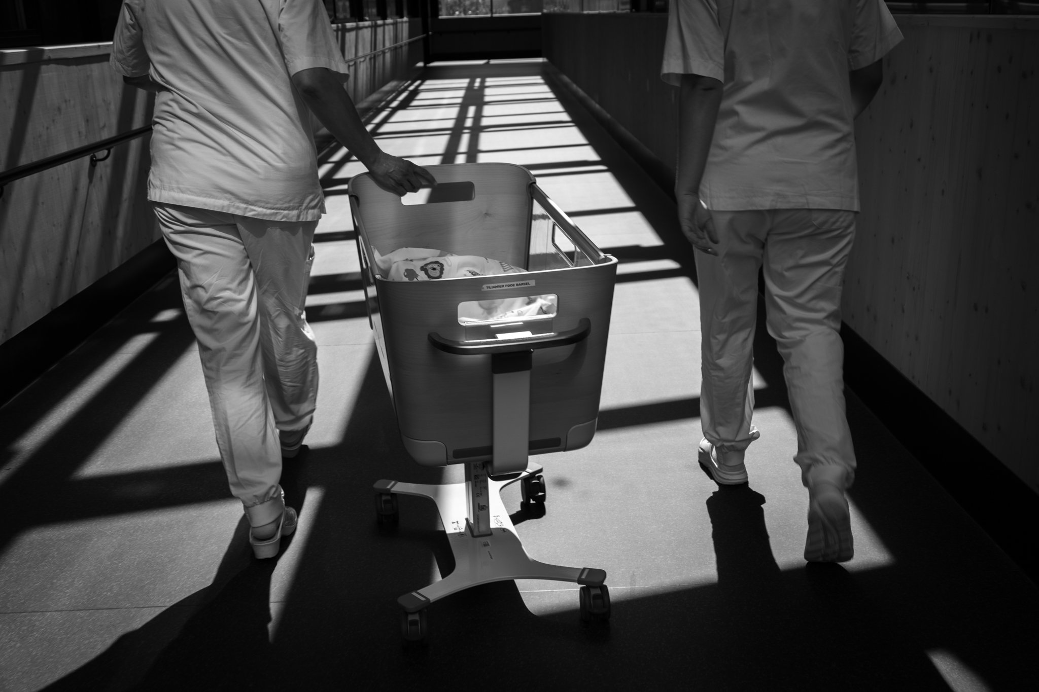 Two healthcare workers dressed in scrubs walking down a hospital corridor pulling a mobile baby incubator on wheels.