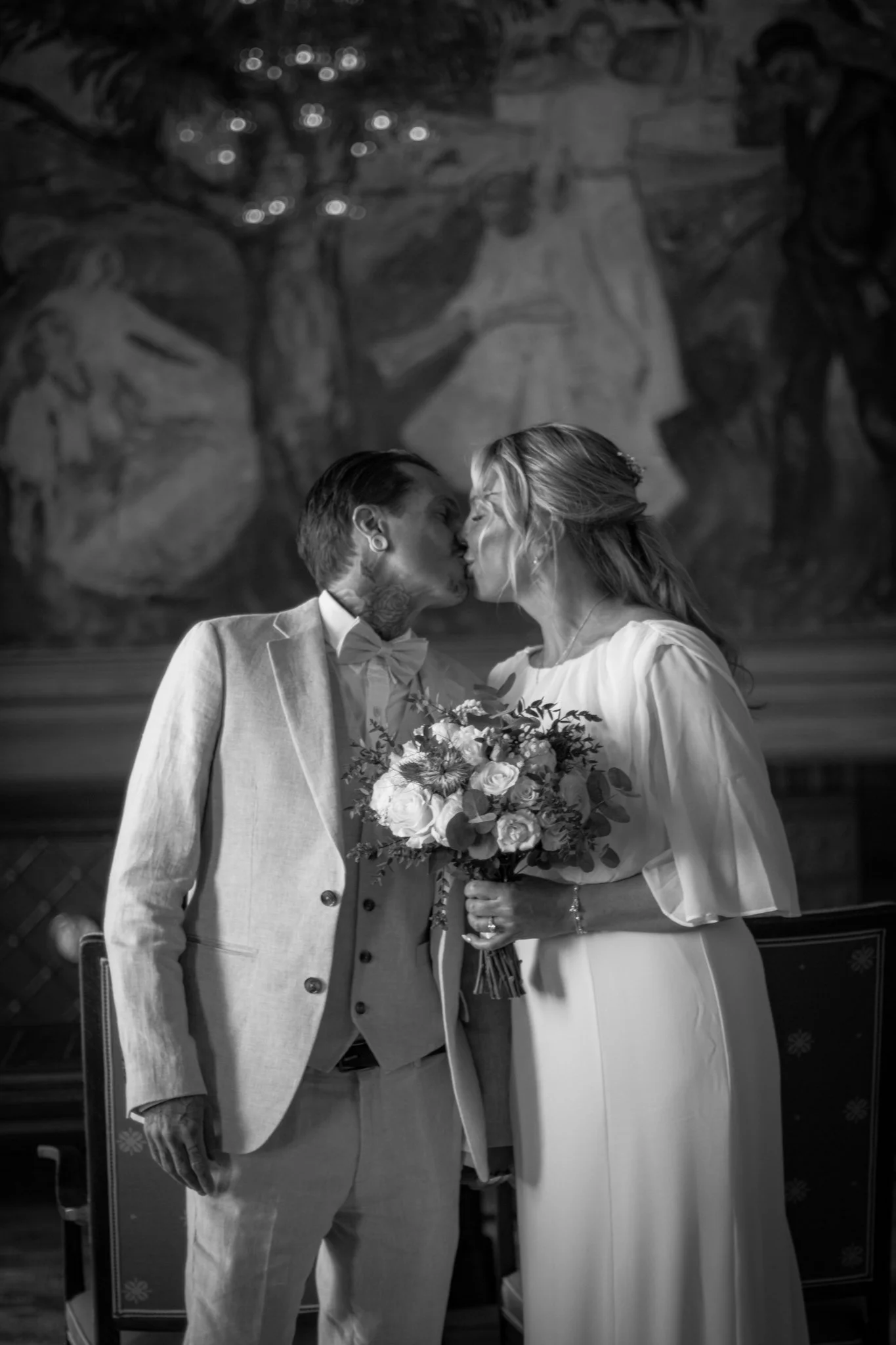 A black and white photo of a couple sharing a kiss, dressed in wedding attire, with a bouquet of flowers in the woman's hand, standing in front of a mural painting.