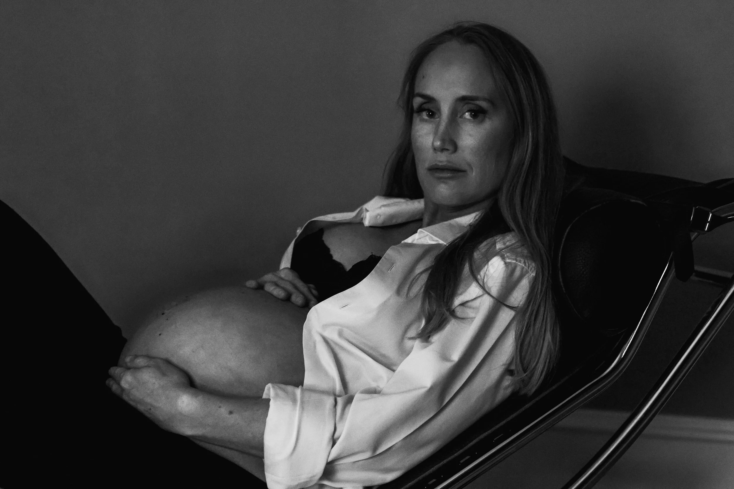 Black and white photo of a woman with long hair sitting in a chair, looking directly at the camera with a serious expression, holding her pregnant belly
