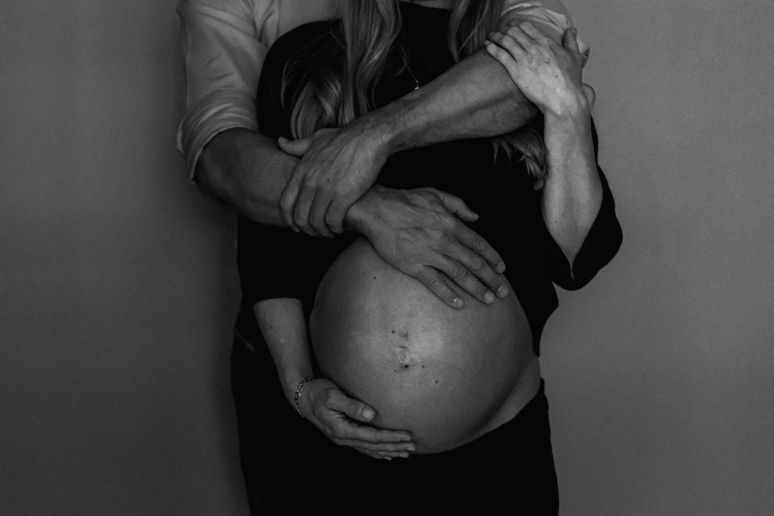 Black and white photo of a pregnant woman being embraced from behind by another person, with their arms around her belly