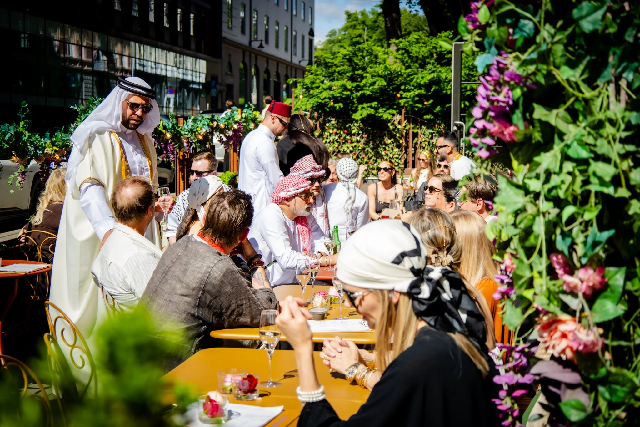 A group of people dressed in Middle Eastern and Mediterranean attire dining outdoors at a restaurant with pink and purple flowers and green trees and buildings in the background.