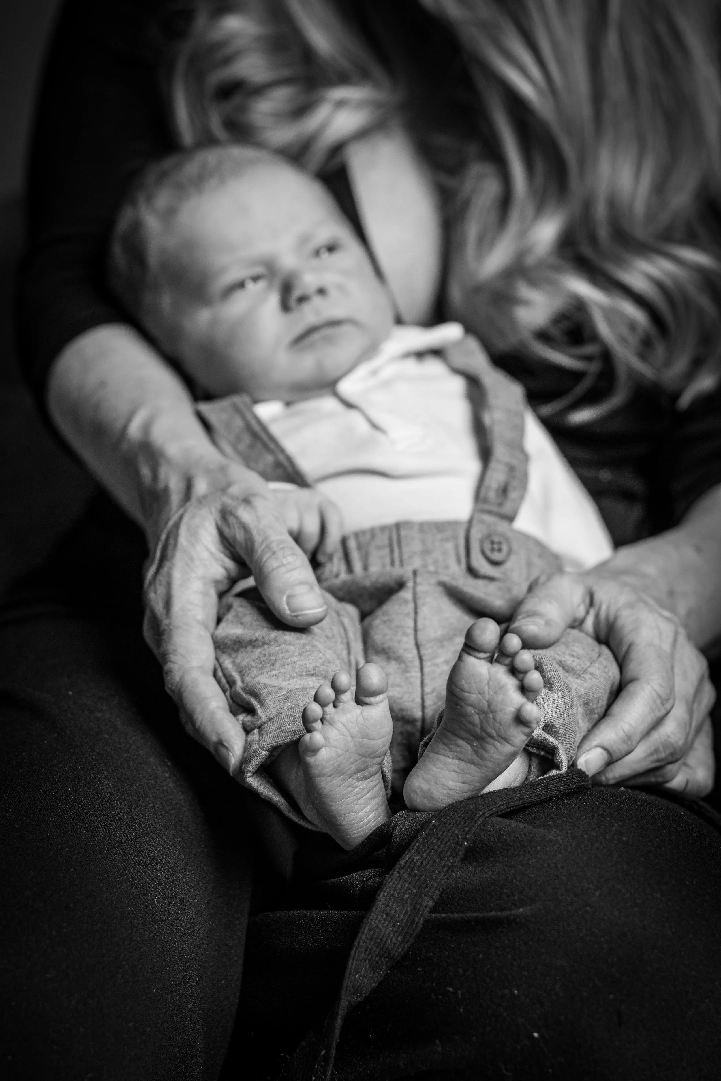 A baby lying on an adult's lap, with the adult's hand gently touching the baby's foot. The baby is looking upwards with a calm expression. The photo is in black and white.