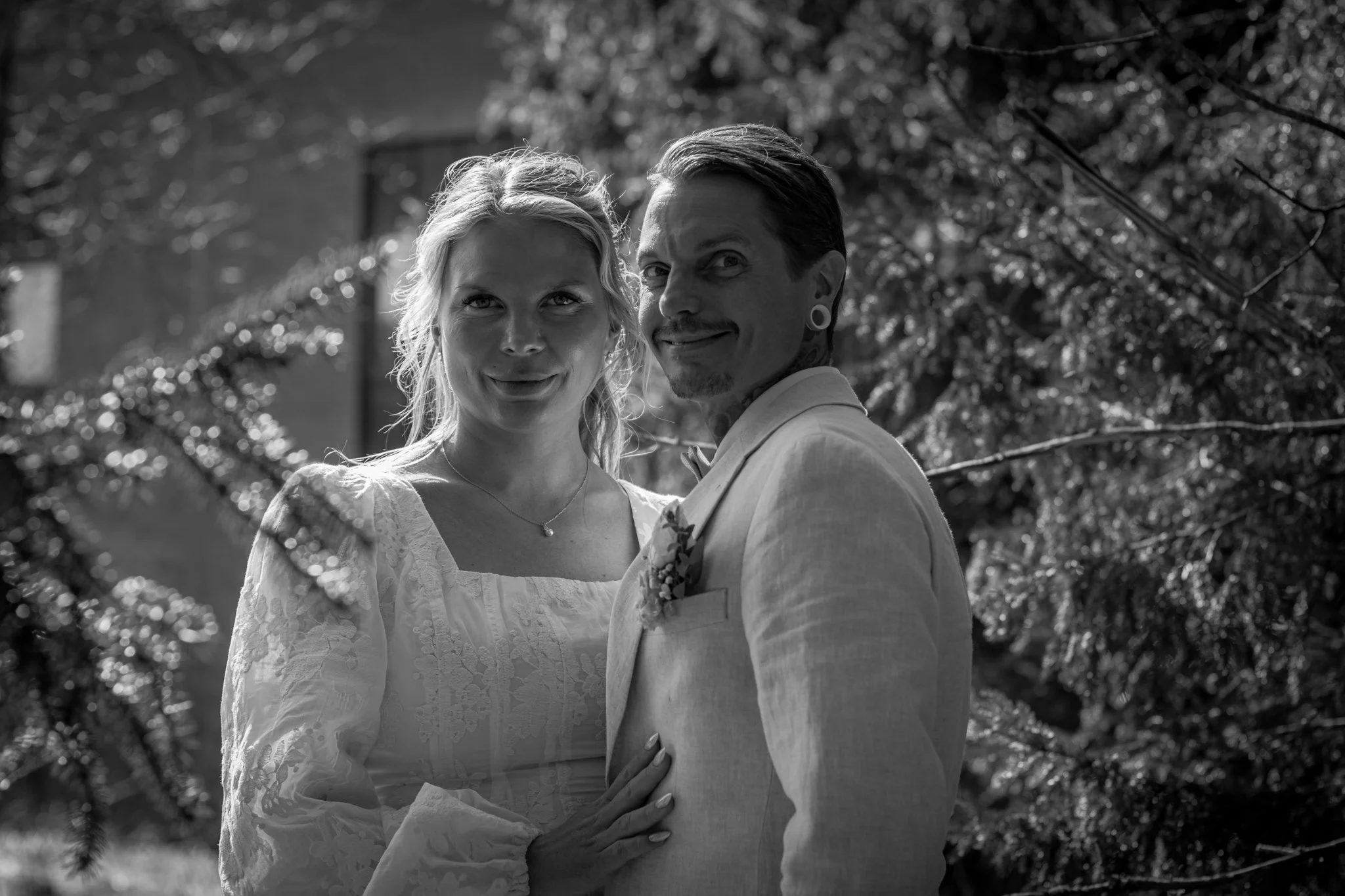 Black and white photo of a couple, the woman in a white lace dress and the man in a light-colored suit, standing outdoors with trees and a building in the background. They are close together, smiling, and looking at the camera.