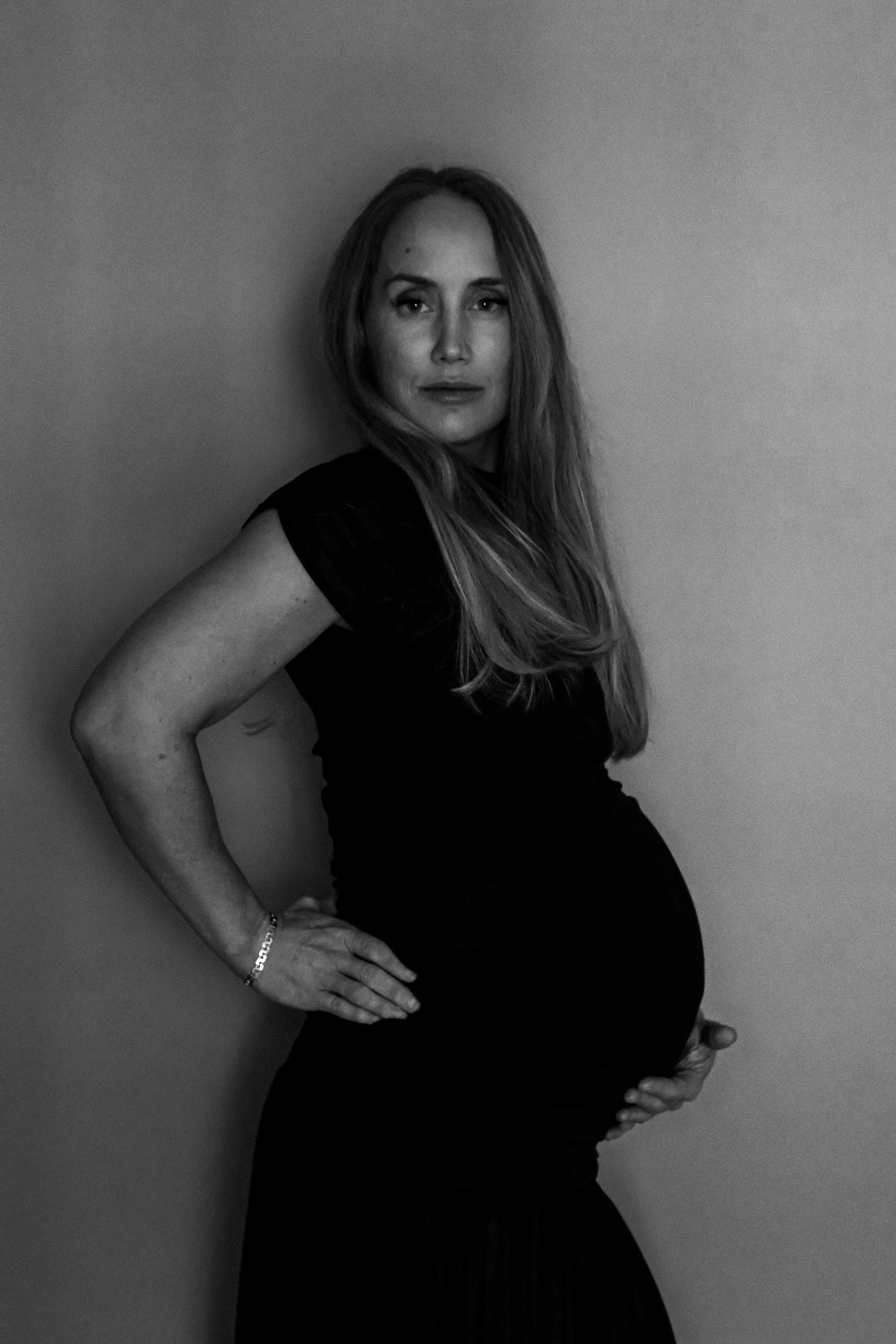 Black and white photograph of a pregnant woman wearing a black dress, standing sideways with one hand on her hip and the other on her belly, against a plain wall.