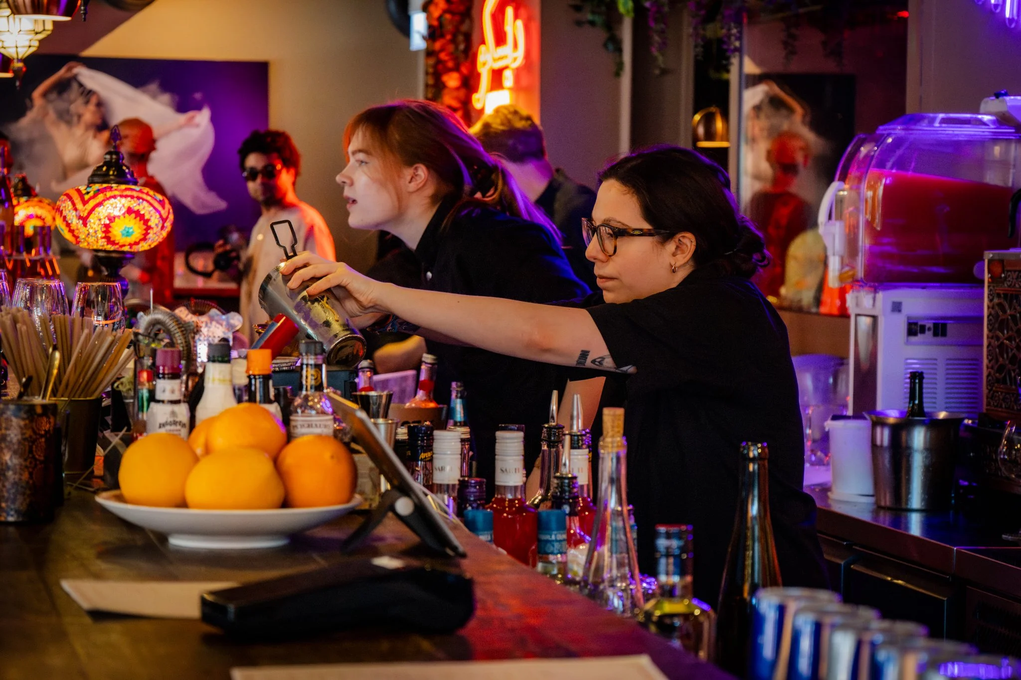Bar scene with two women preparing drinks, colorful bottles and oranges on the counter, vibrant neon and decorative lighting, and other patrons in the background.