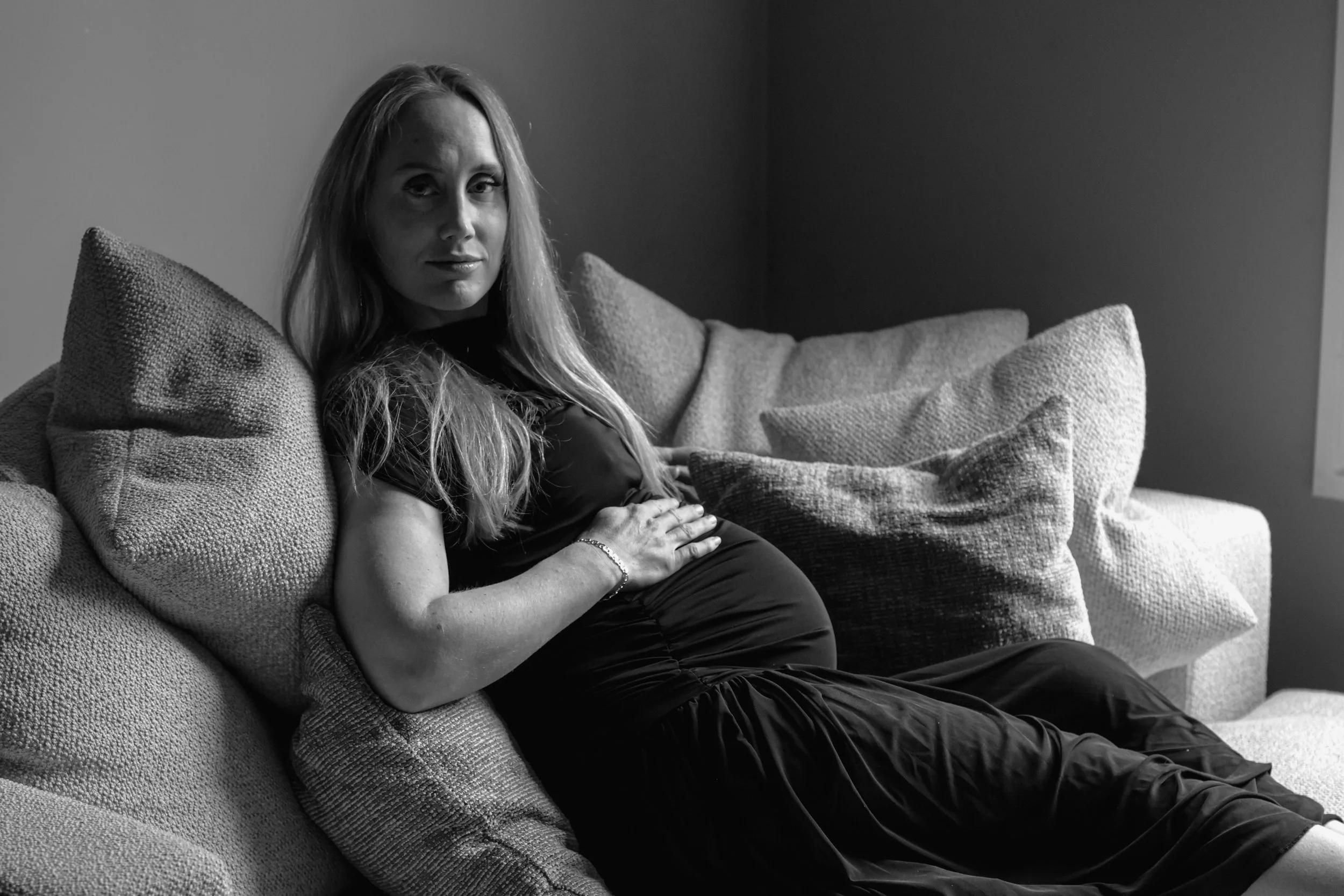 A woman with long hair sitting on a sofa holding her pregnant belly, looking at the camera in a black and white photo.