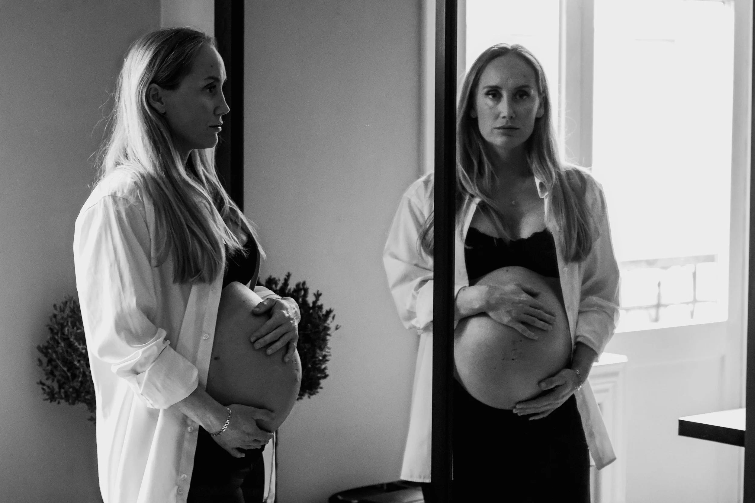 Pregnant woman looking into a mirror and the reflection looking back. Holding her pregnant belly.