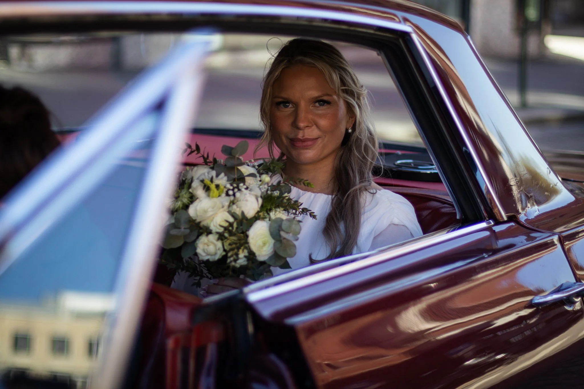 A woman sitting inside a vintage car holding a bouquet of white and green flowers, viewed through the car window.