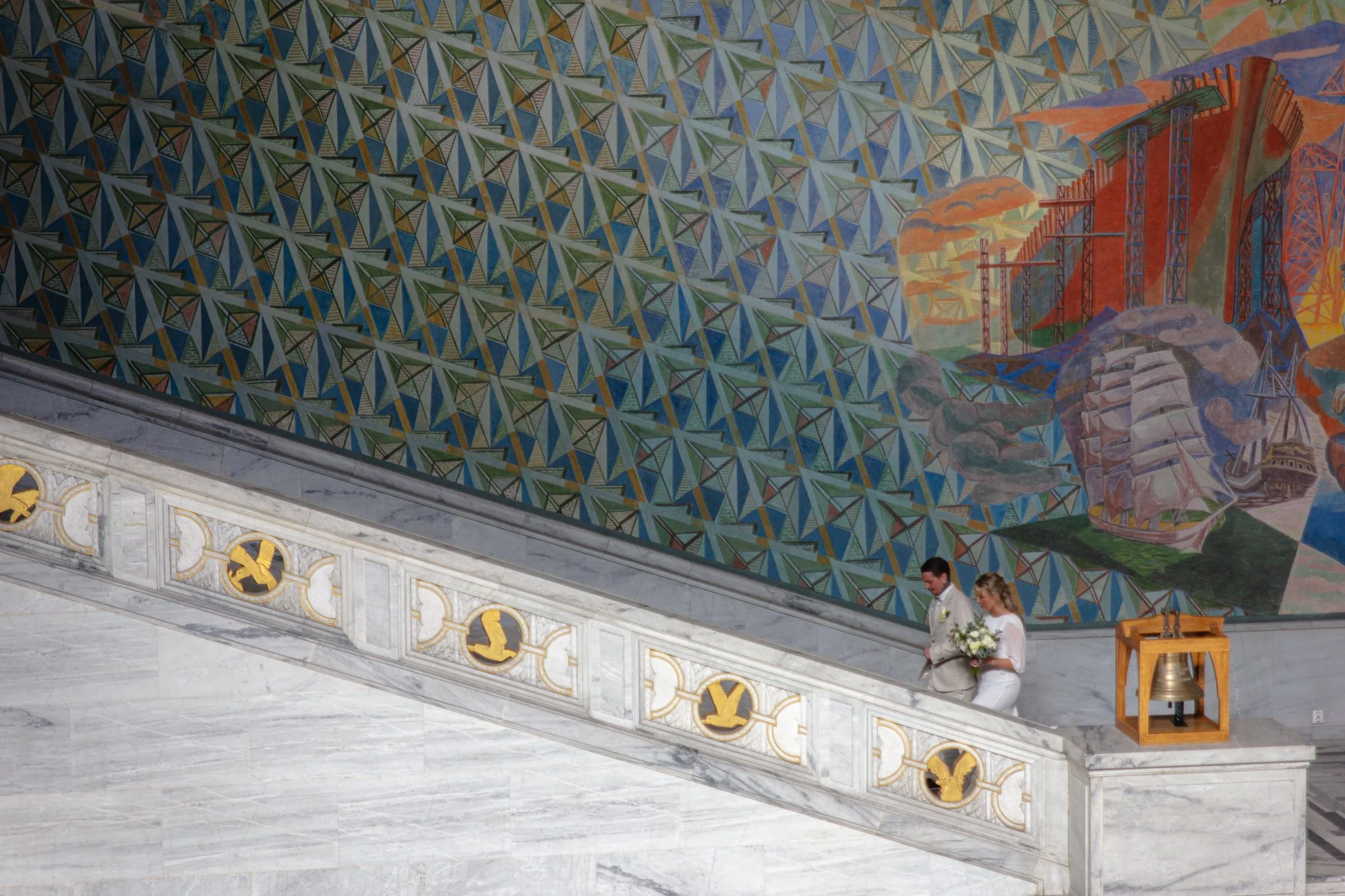 A religious wedding ceremony inside a church with a colorful geometric mural on the ceiling, marble walls, and two people standing on the stairs, one of whom is holding a bouquet of flowers.