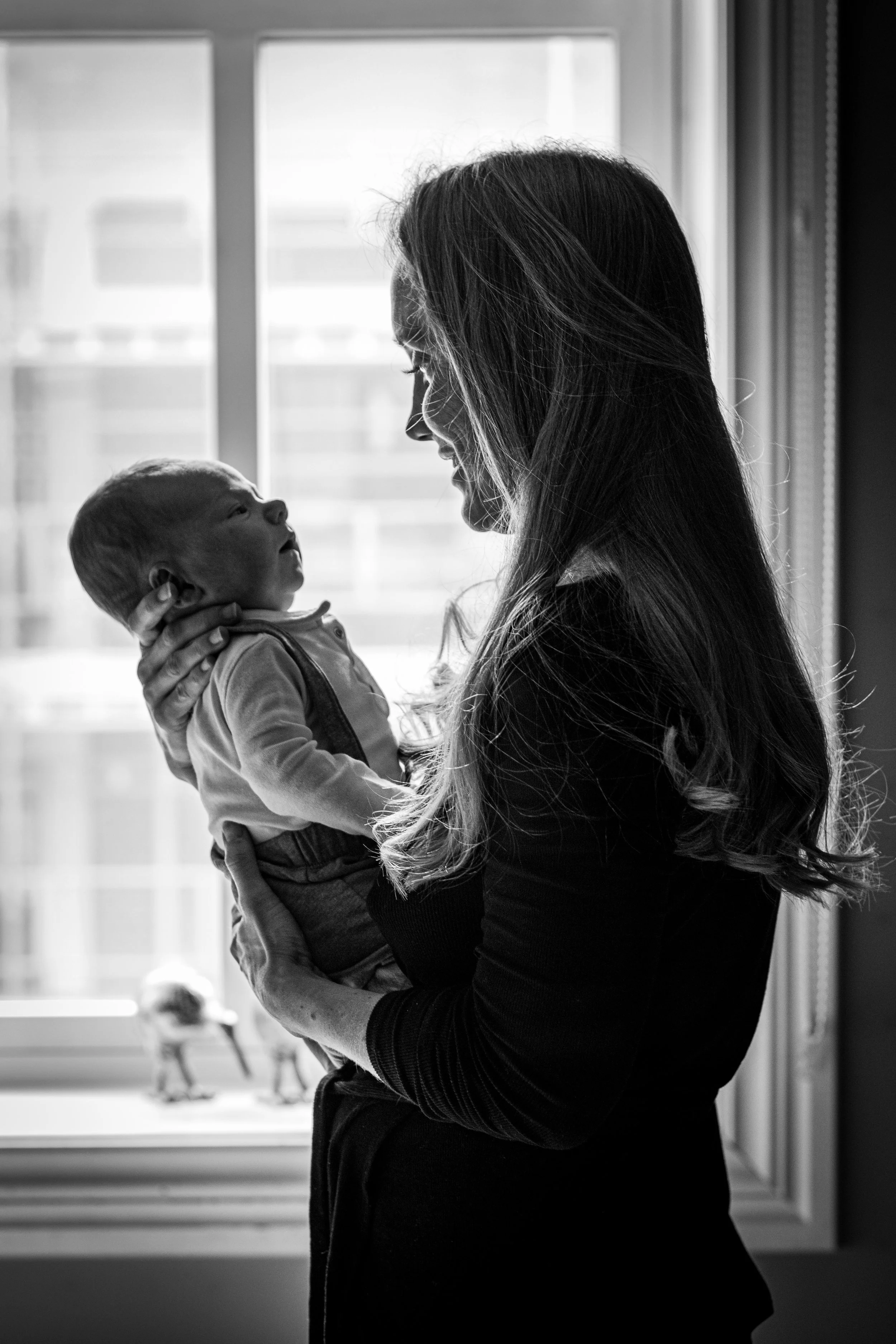 A woman holding a baby in front of a window, with the woman smiling and the baby looking up at her, in black and white.