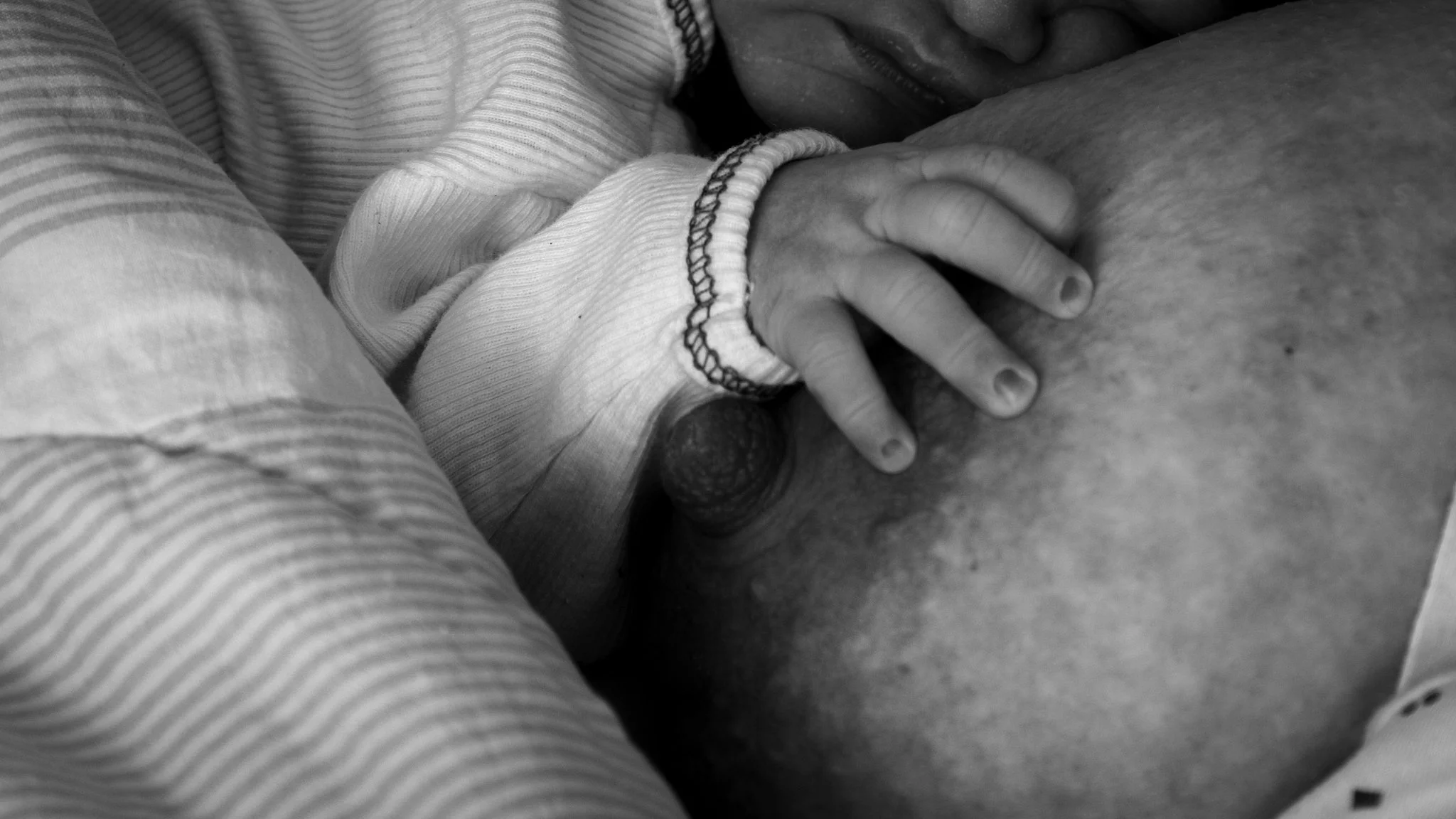 A person lying down with a child snuggling close, hand on the child's back, baby holding the woman's breast, in black and white
