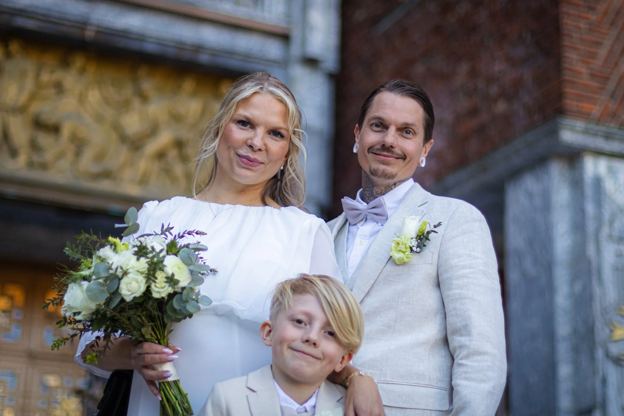 A happy bride and groom with two children, standing outdoors in front of brick buildings, during a wedding celebration. The bride holds a bouquet of flowers, and the couple is dressed in formal attire.