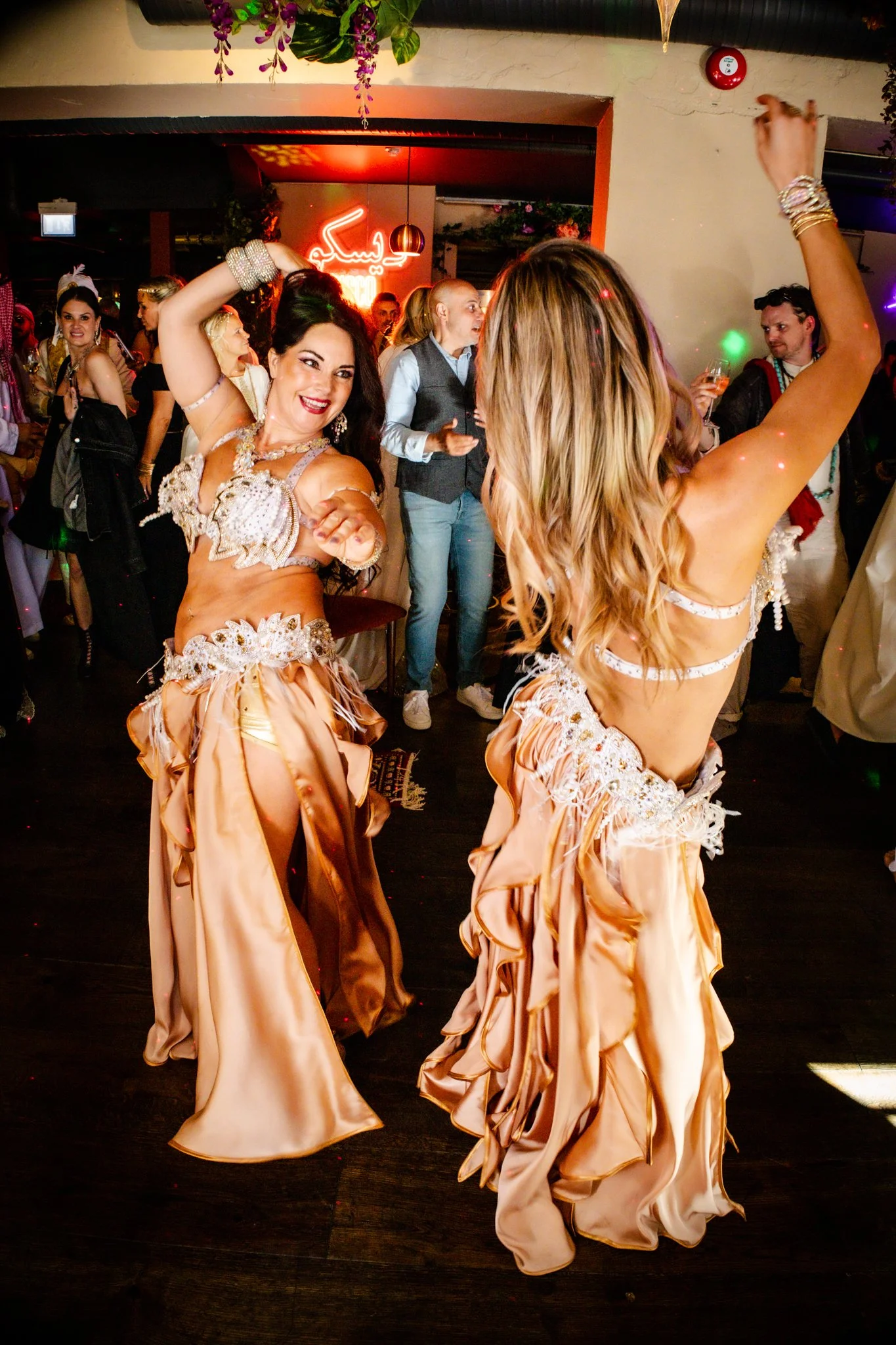 Two women in elaborate belly dance costumes performing at a lively celebration or party, with one woman smiling and dancing, and a crowd observing in the background.