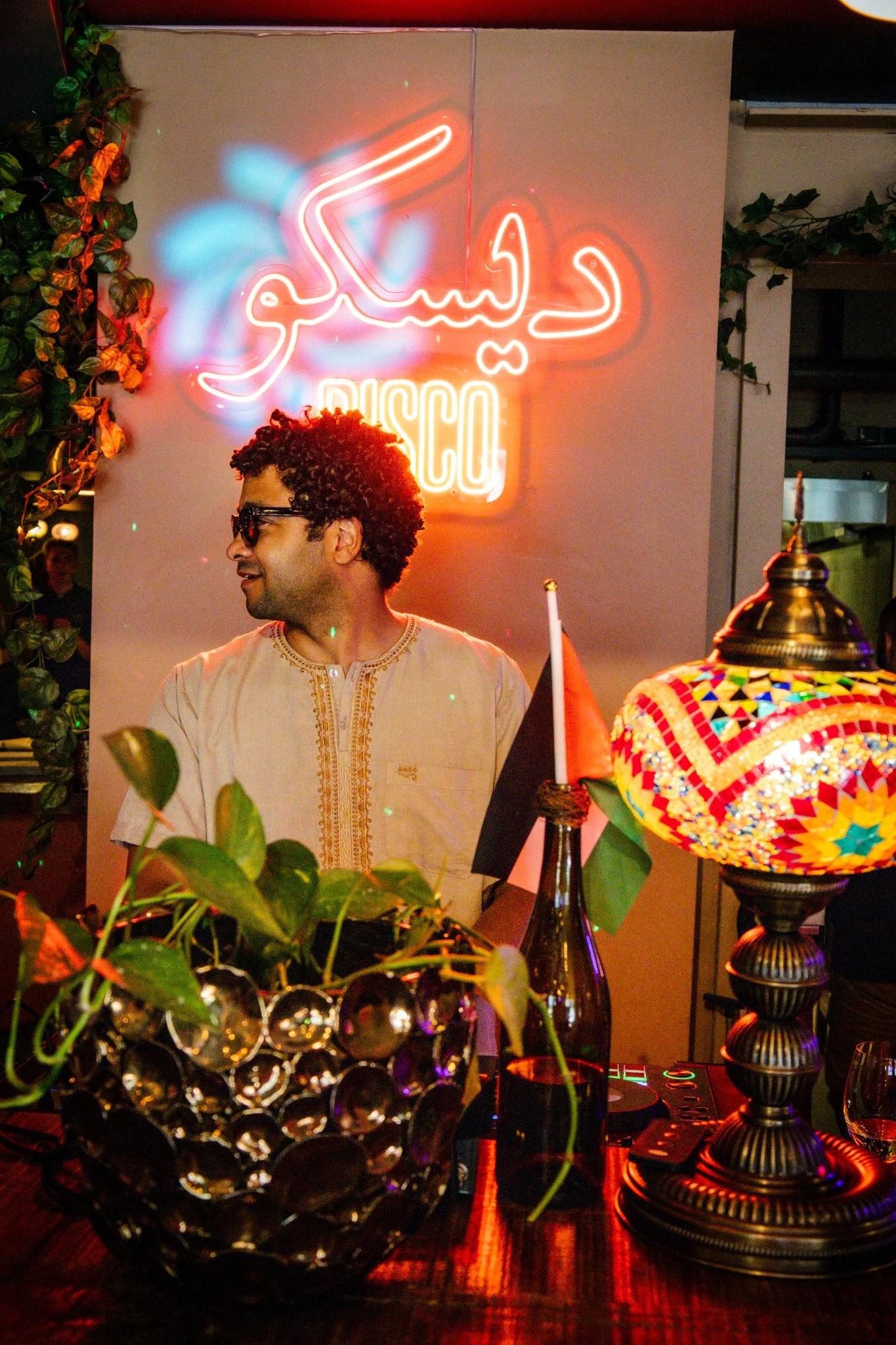 A man with curly hair and sunglasses smiling at a party with colorful decor, including a neon sign in Arabic and a decorative lamp, surrounded by plants and bottles on the table.