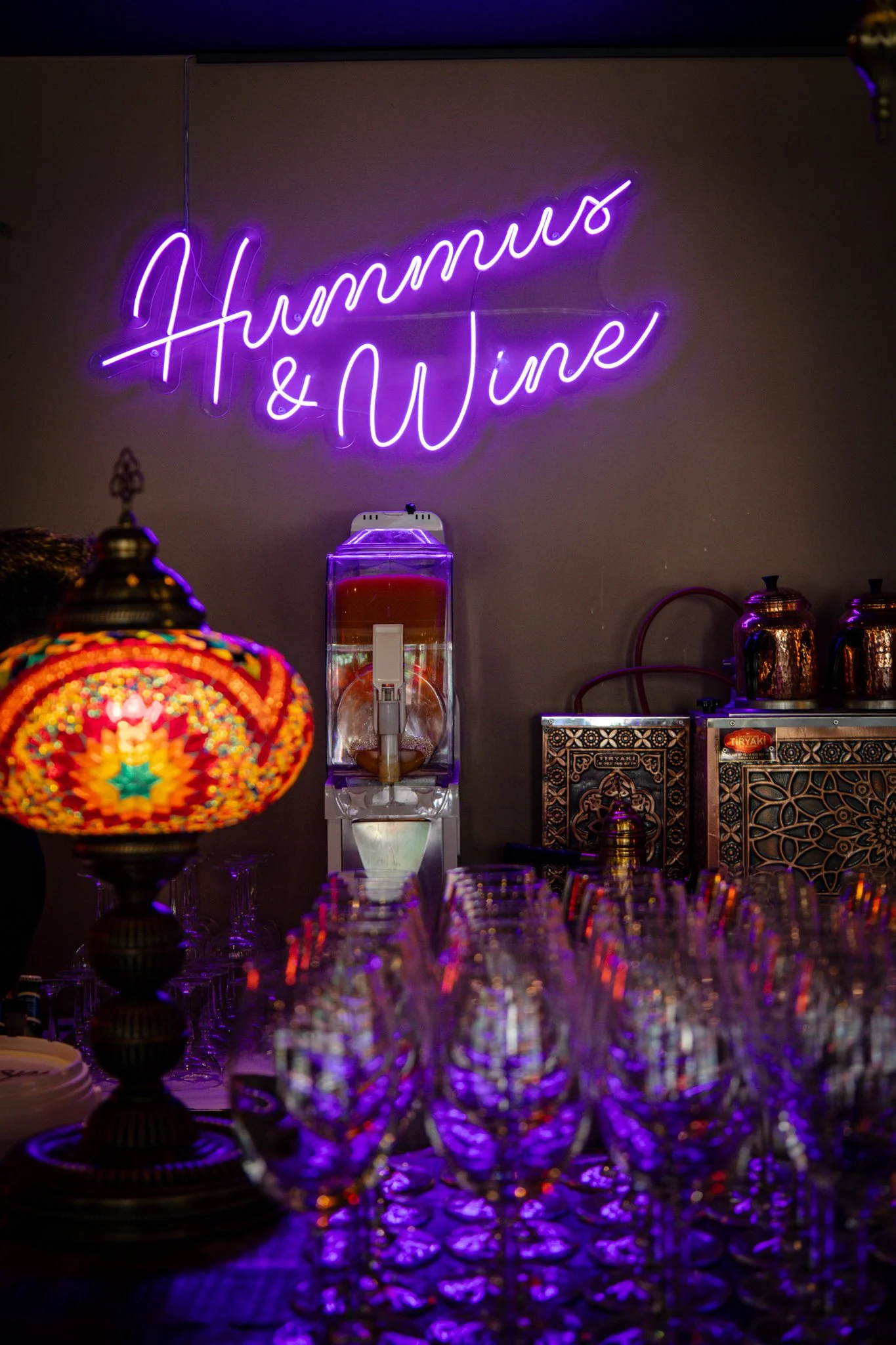Neon sign with purple cursive writing saying 'Hummus & Wine' on a beige wall in a dimly lit setting with glasses and decorative items on the table.