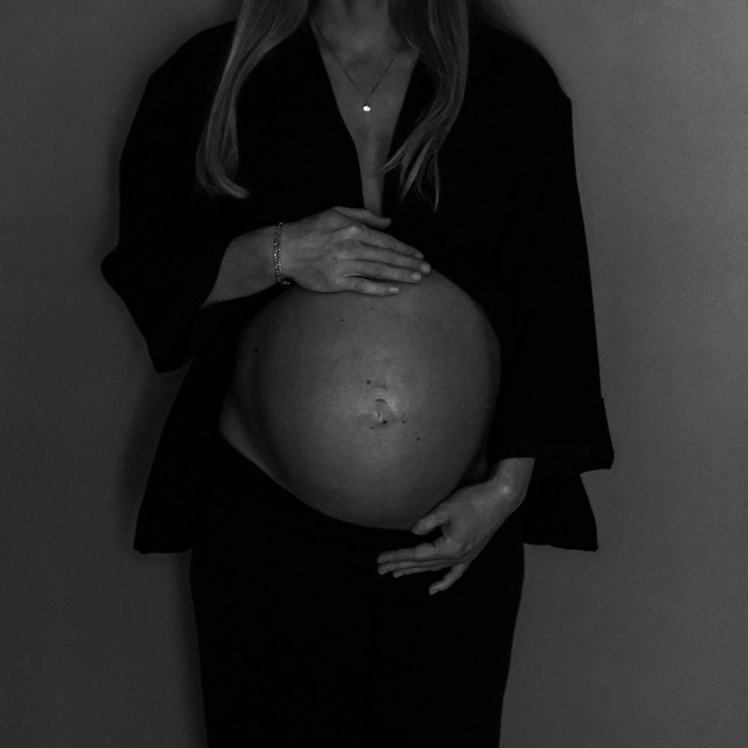 Black and white image of a woman with long hair, wearing a necklace and a bracelet, dressed in a black top, standing with her arms holding her pregnant belly, in a dimly lit setting