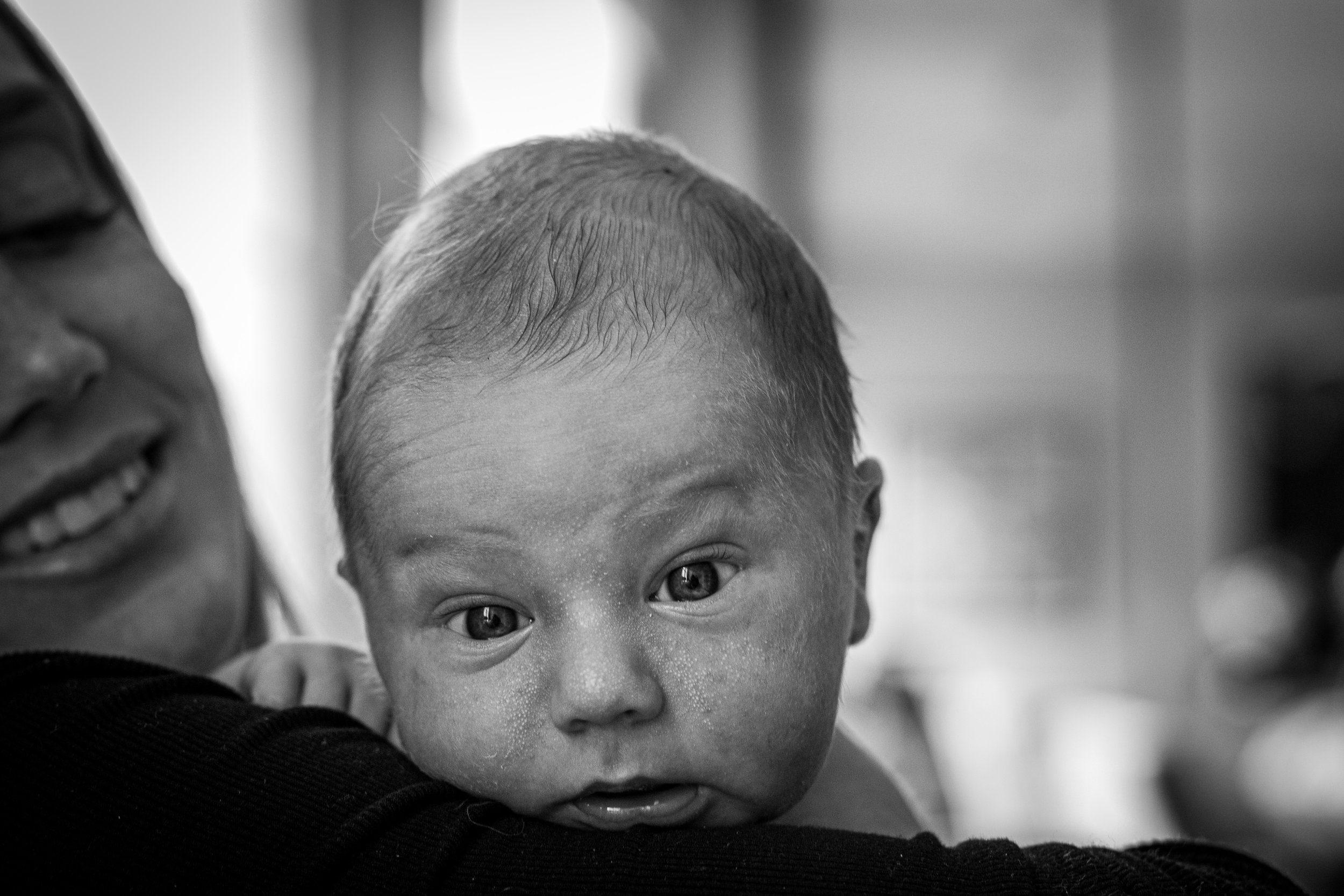 A newborn with light skin and short hair looks directly at the camera while being held by his mother wearing a dark shirt, with a background of blurred indoor setting.