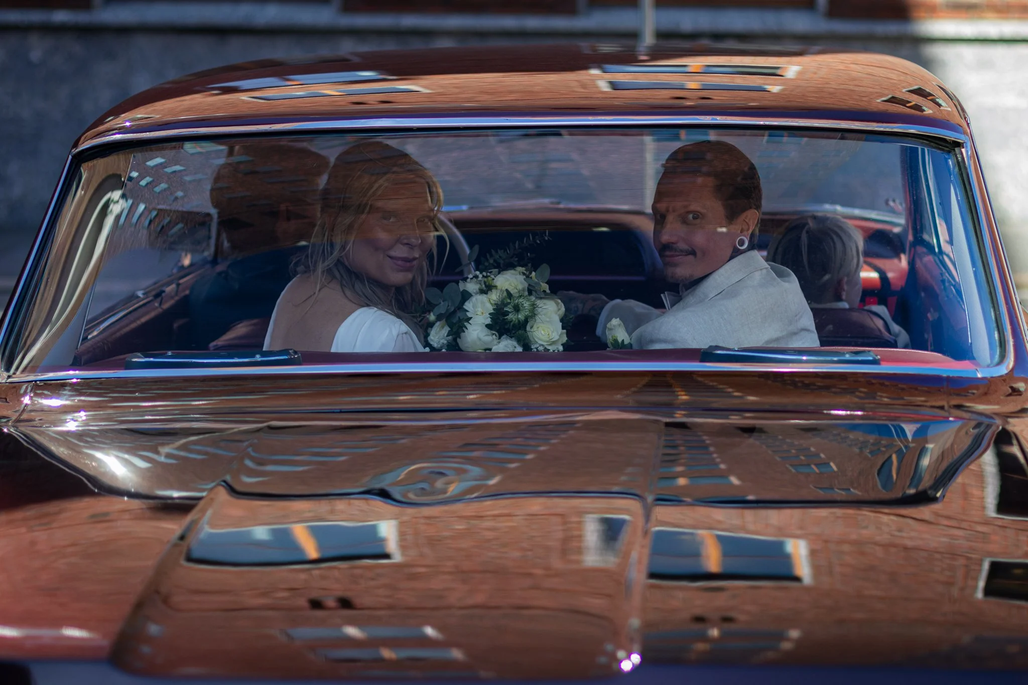 A woman and a man sitting inside a vintage brown car, looking back and smiling, with the woman holding a bouquet of white flowers, through the windshield reflecting buildings.