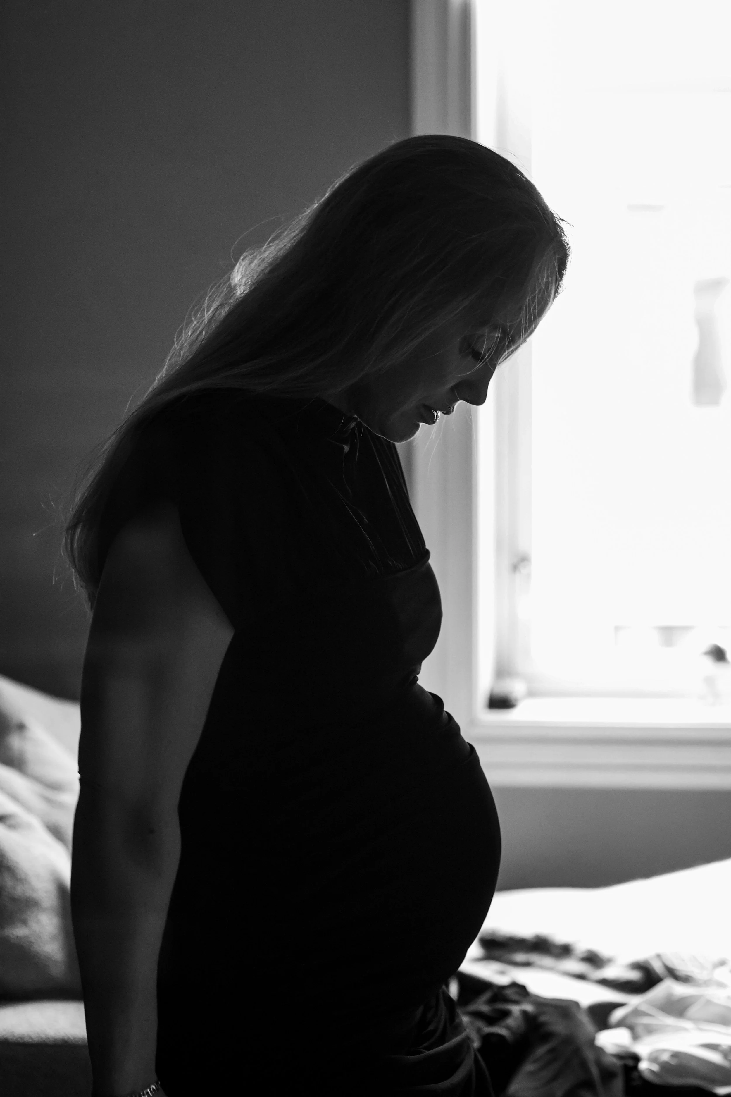 Black and white side profile of a pregnant woman standing by a window with natural light streaming in.