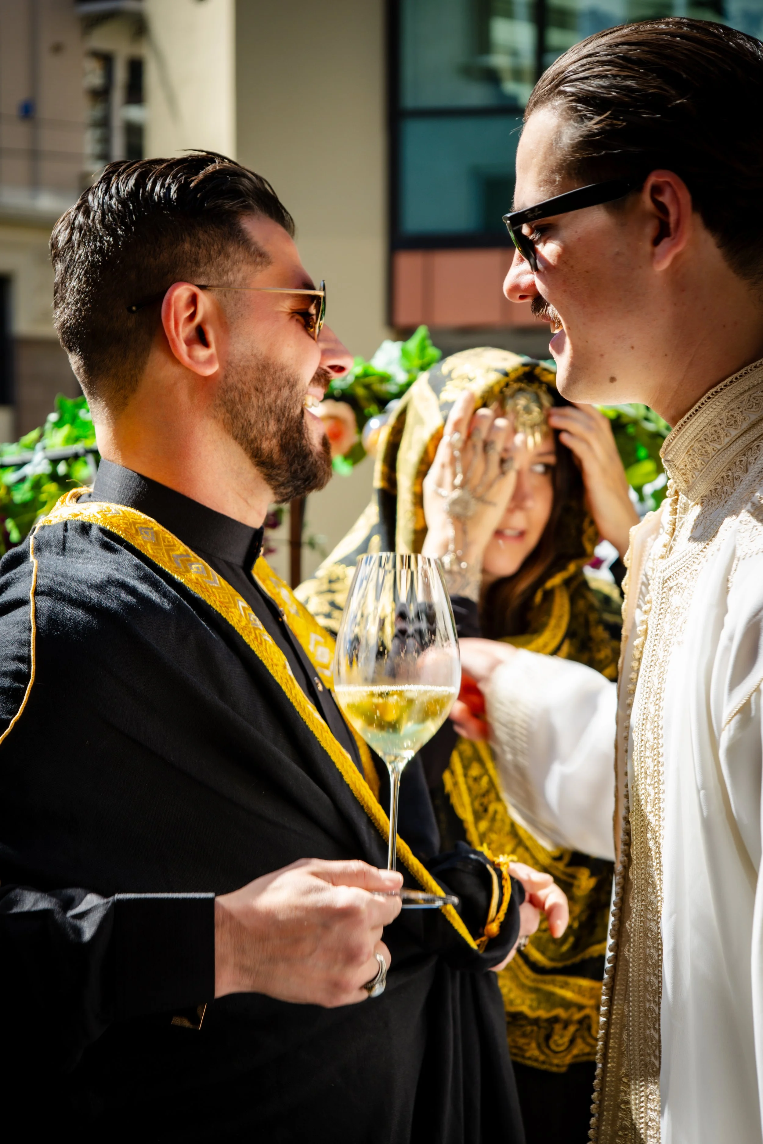 Two men laughging and chatting at a party, champagne in hand. A woman in the background observing
