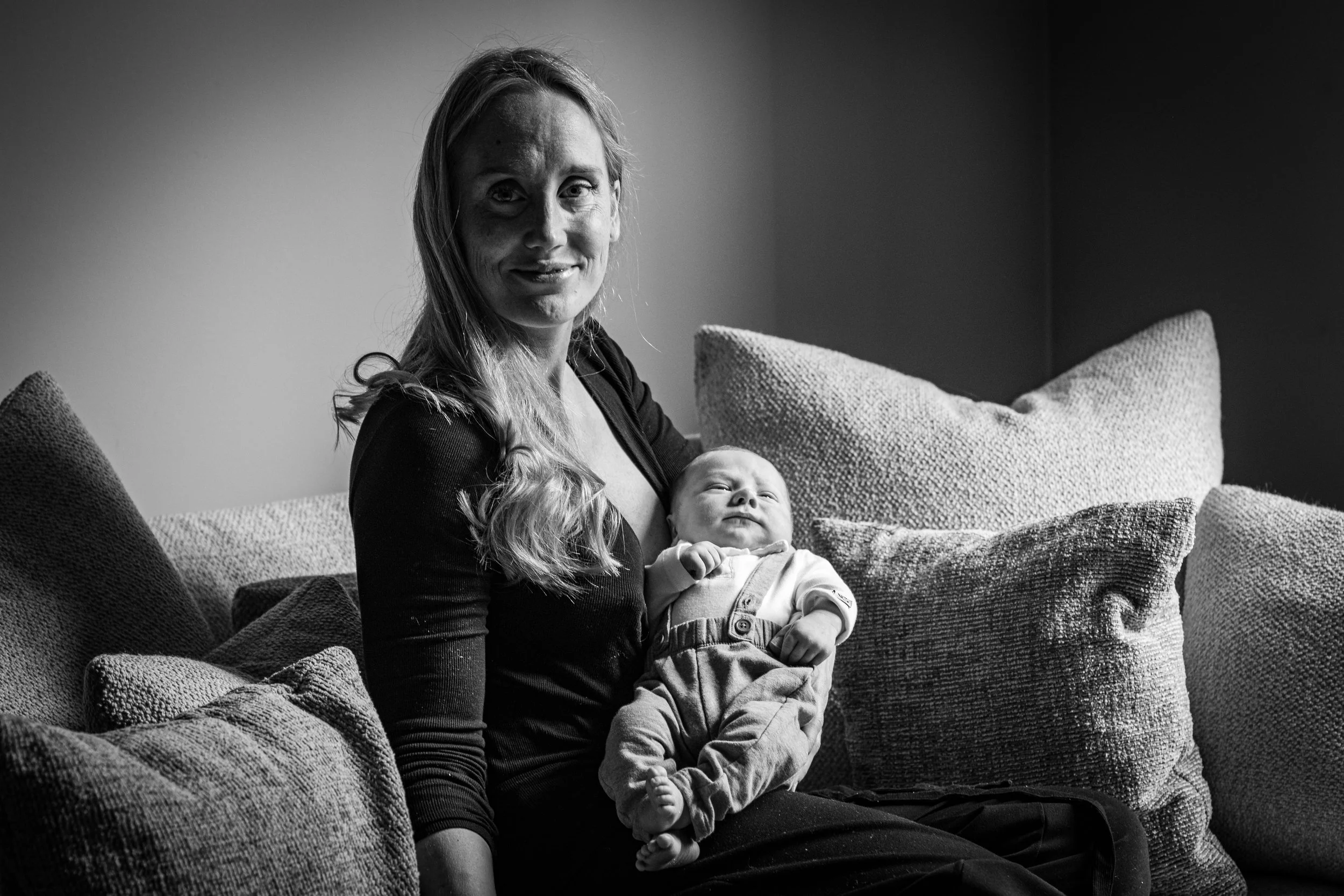A woman with long hair smiling at the camera while holding a baby, sitting on a sofa with multiple textured cushions in a room with plain walls.
