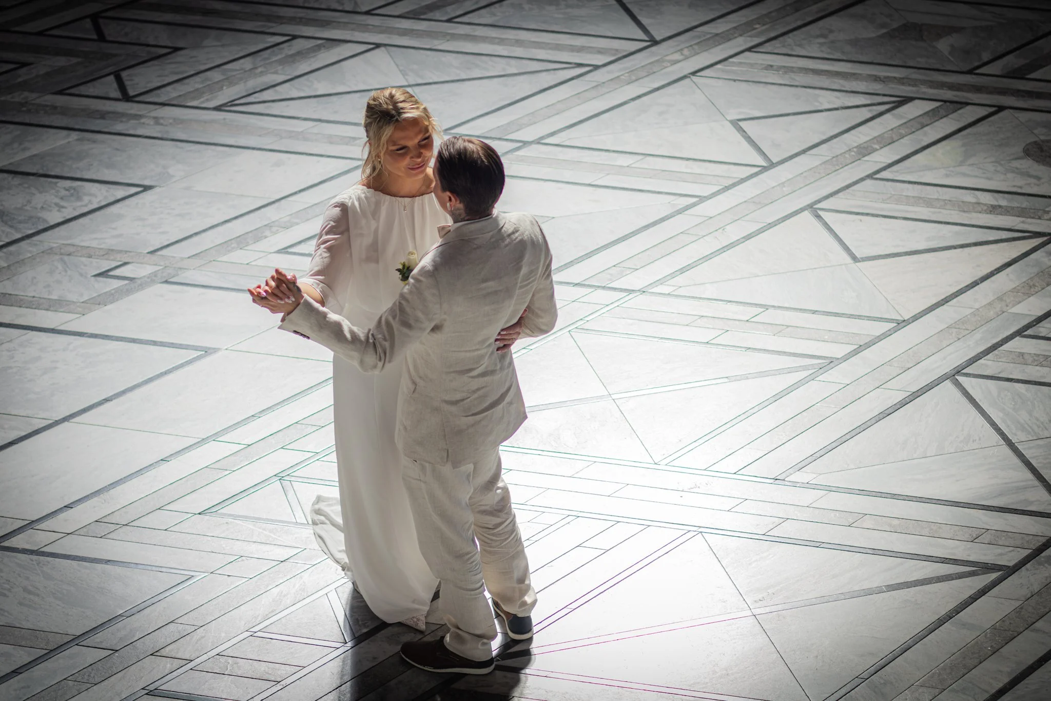 A couple dancing together indoors on a marble floor, with the woman wearing a white dress and the man dressed in a light suit.