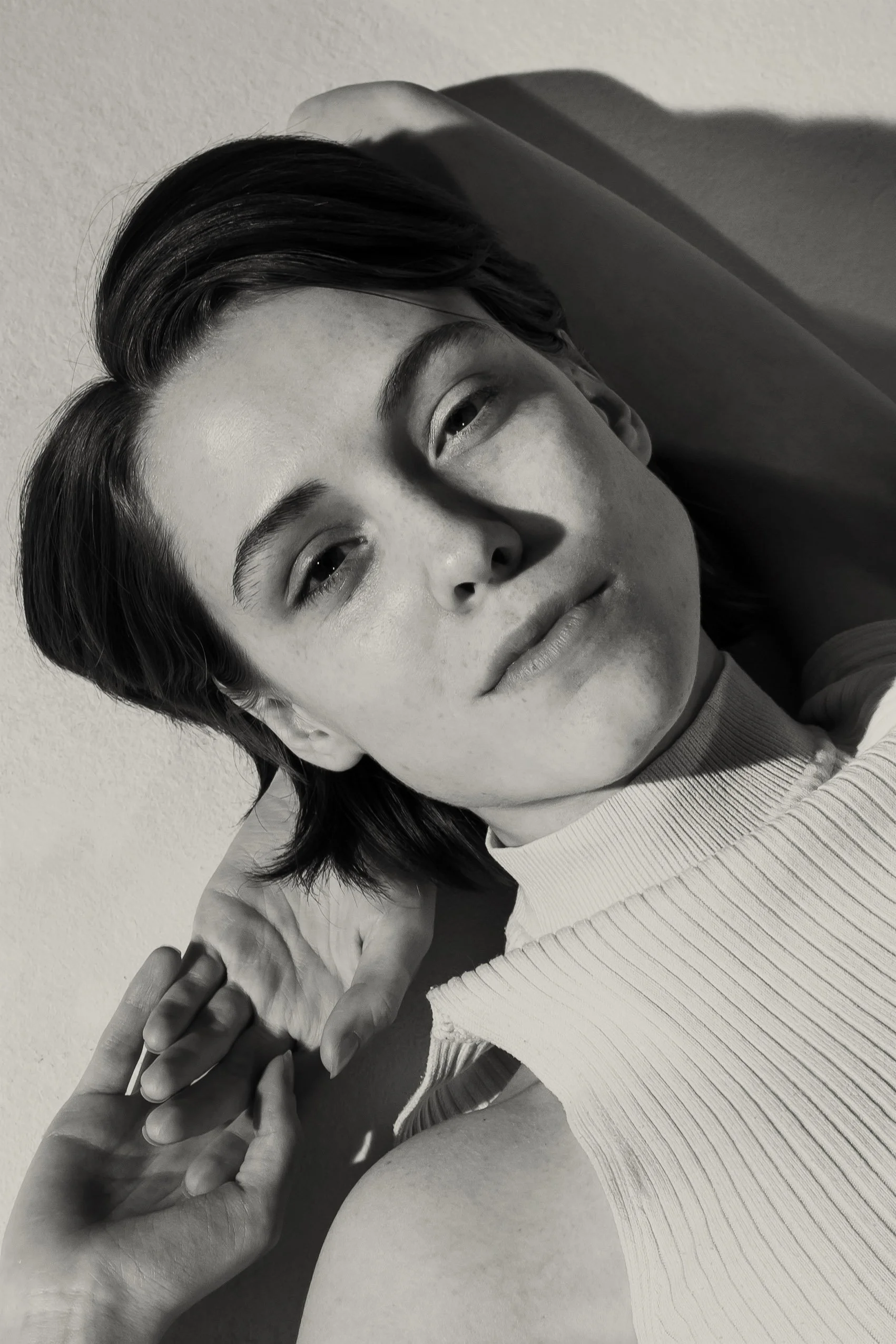 A black-and-white photo of a woman with short dark hair, wearing a ribbed tank top, posing with her hand near her face, against a plain wall background.