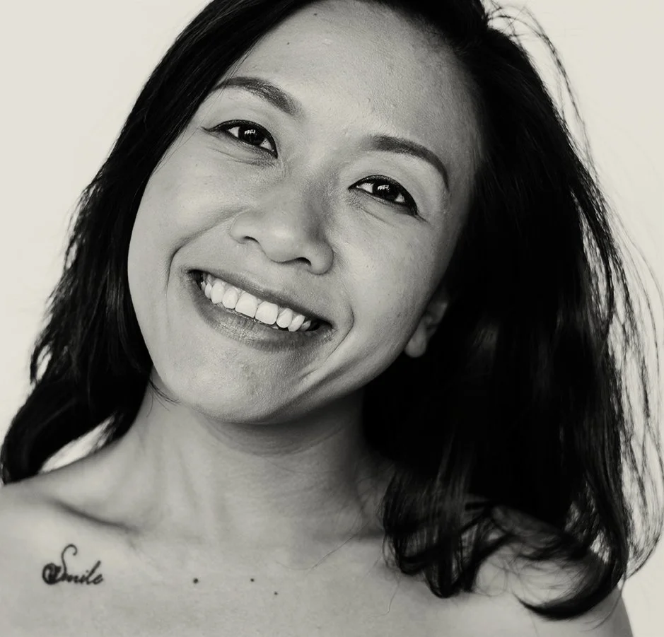 Black and white portrait of a woman smiling showing her teeth, with shoulder-length dark hair and a tattoo on her collarbone that says 'Smile'.