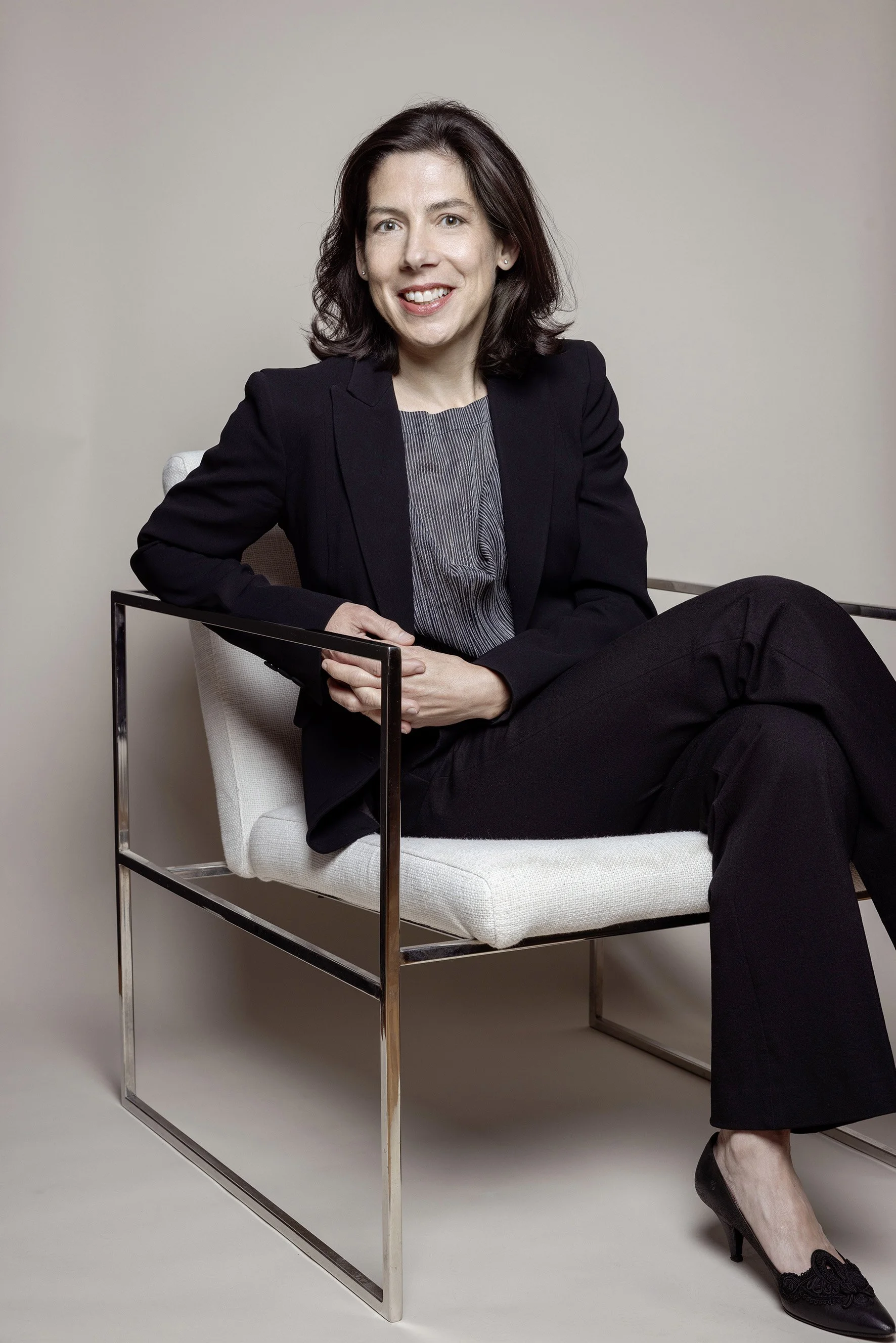 Dr Sandra Handlbauer-Zrust, wearing a black blazer, gray blouse, and black pants, sitting on a modern chair.