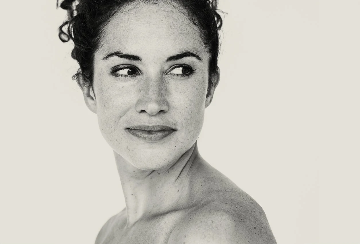 A close-up portrait of a woman with short, curly hair, looking to the side, with freckles, against a plain light background.