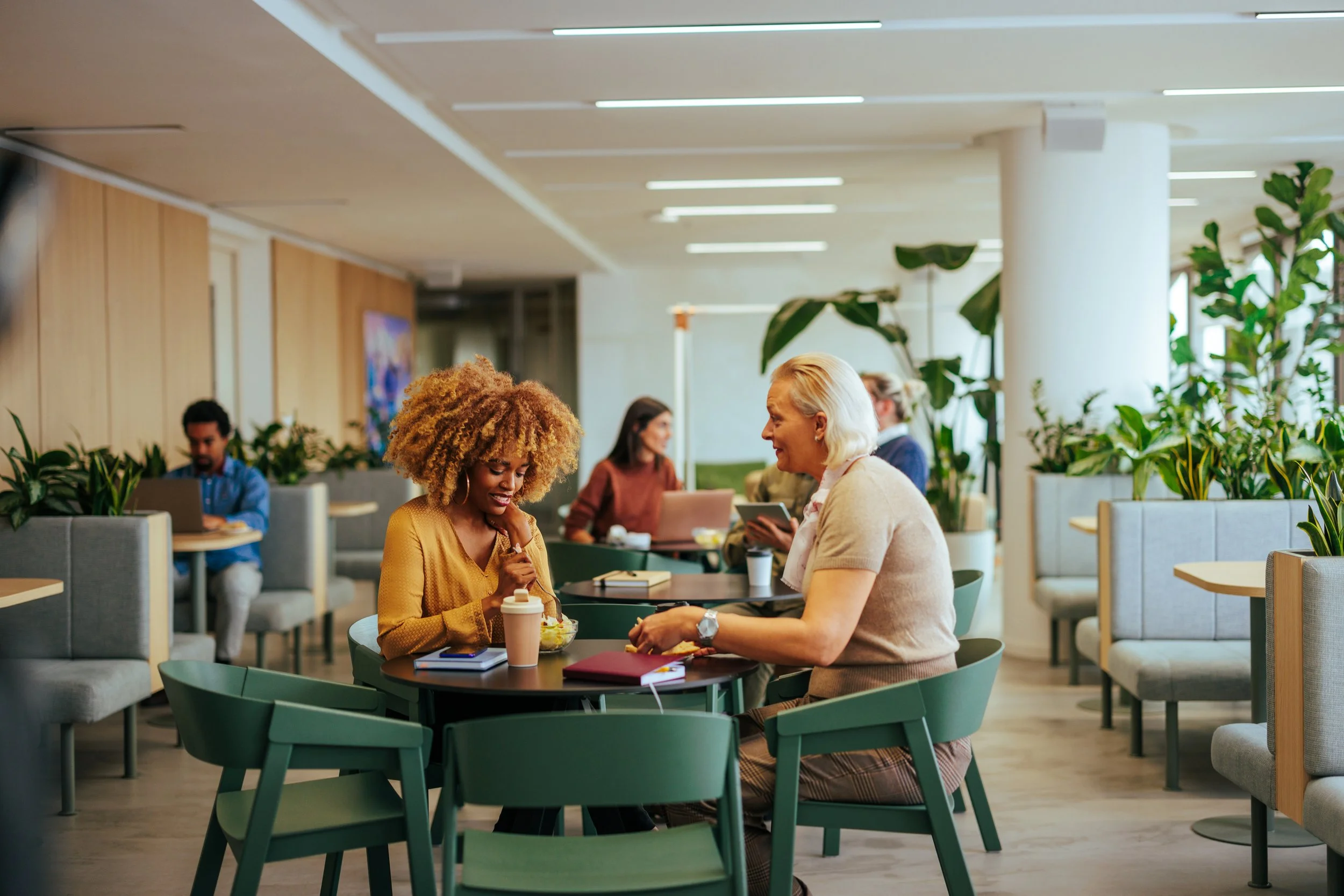 People sitting at tables in a modern, brightly lit café with green chairs and abundant indoor plants, engaging in conversation and work.