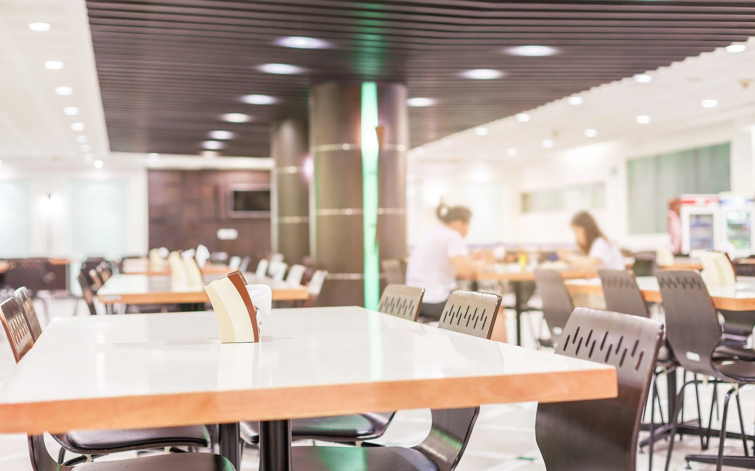 A spacious, well-lit cafeteria with multiple tables and chairs, two people sitting at a table, and a vending machine in the background.