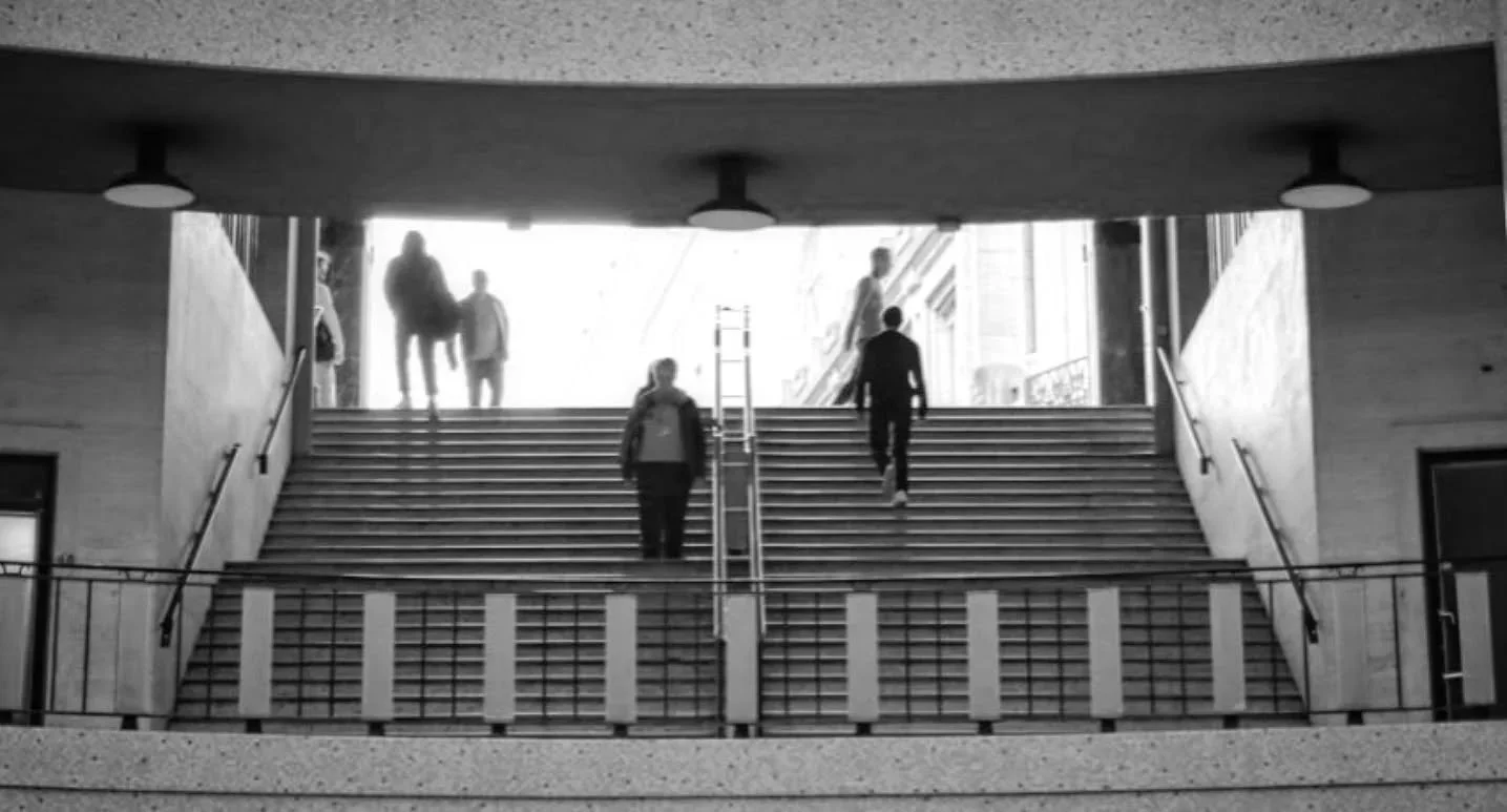 People walking up and down a staircase in a modern building, with some passing through an illuminated entrance or exit.