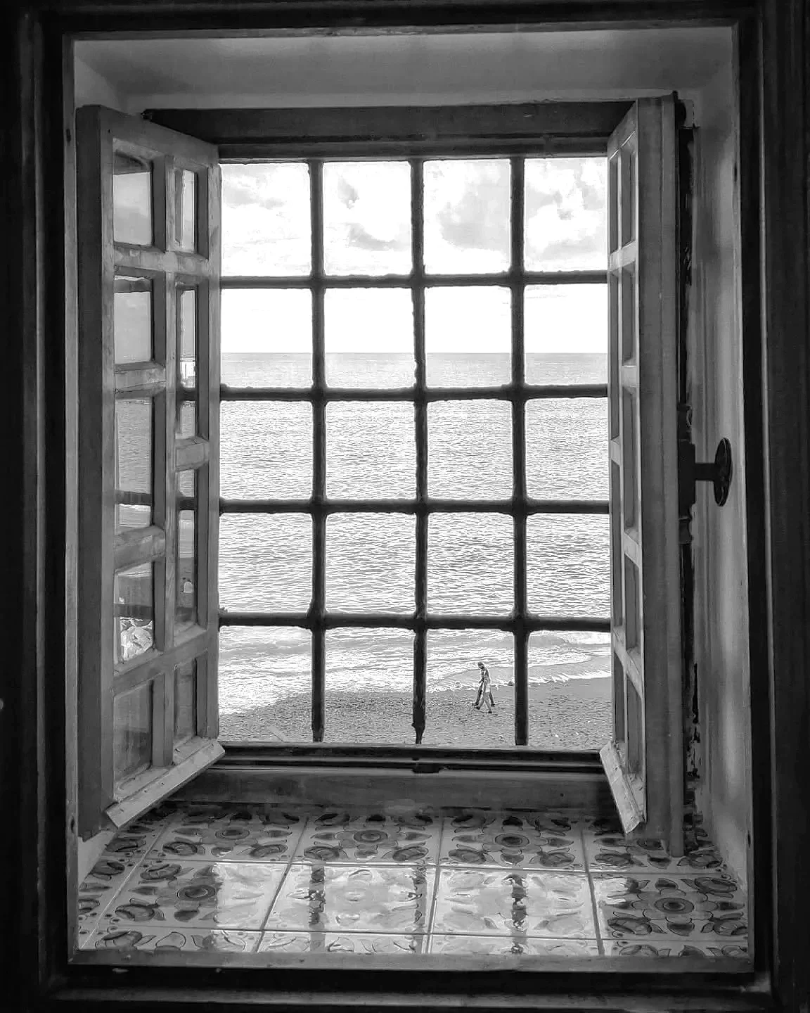 View of a beach through an open window with wooden shutters, overlooking the ocean with a person walking along the shoreline.