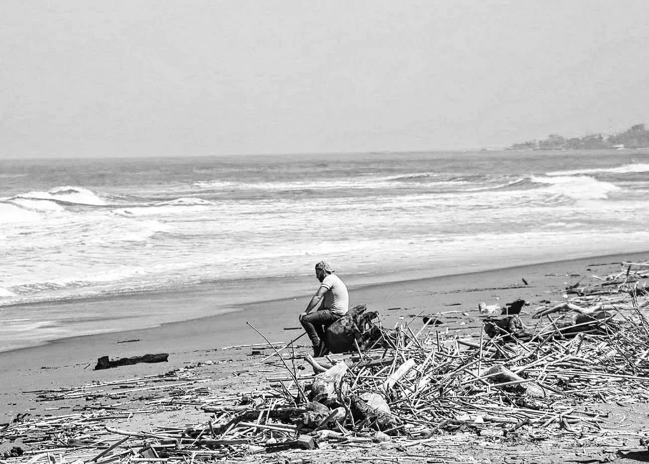 black and white photograph of a solitary figure sitting by the sea, surrounded by shoreline, horizon and traces left by the water.