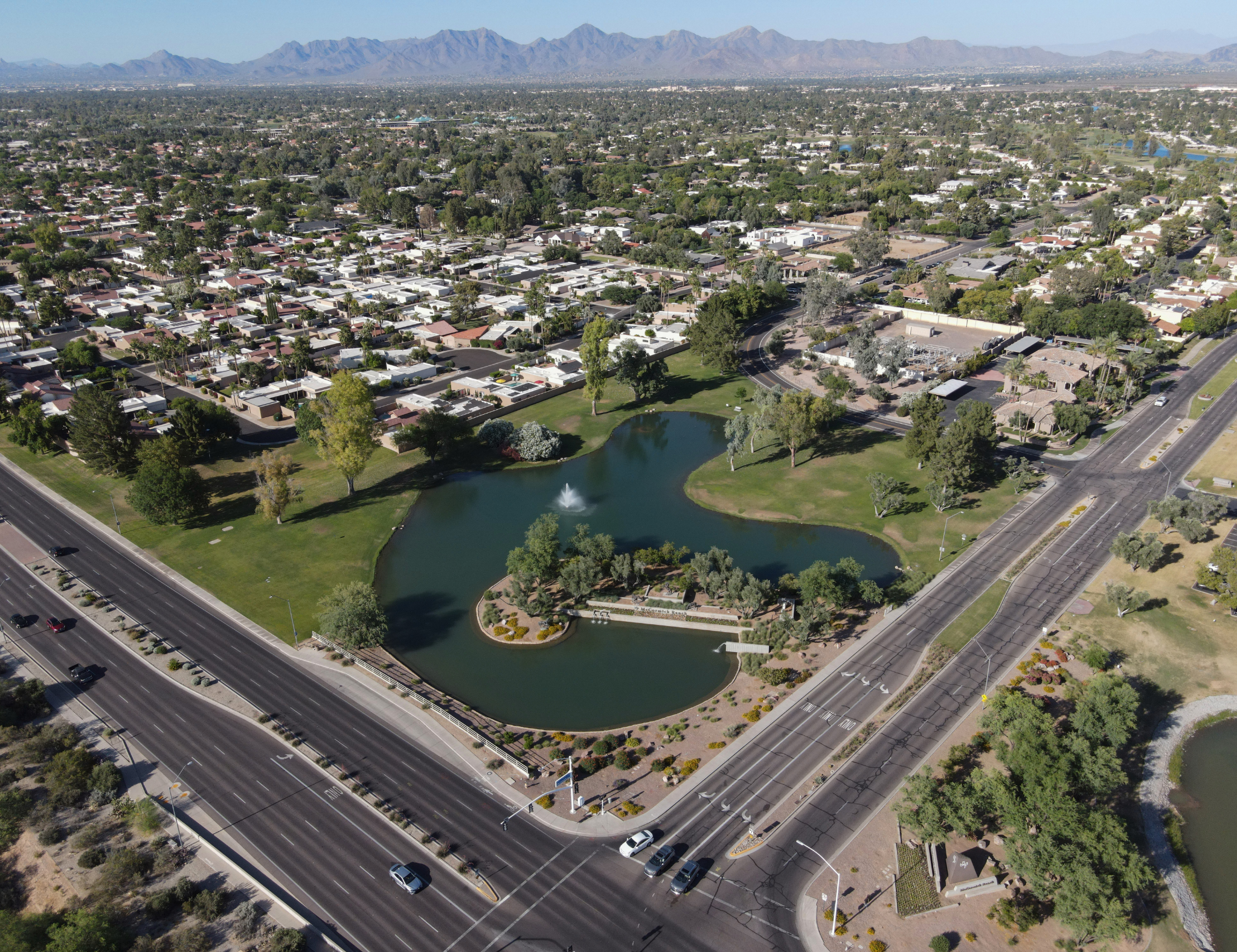 Aerial view of a suburban neighborhood with rows of houses, green trees, a small pond with a fountain, and a highway with cars in the foreground. Mountain range in the background.